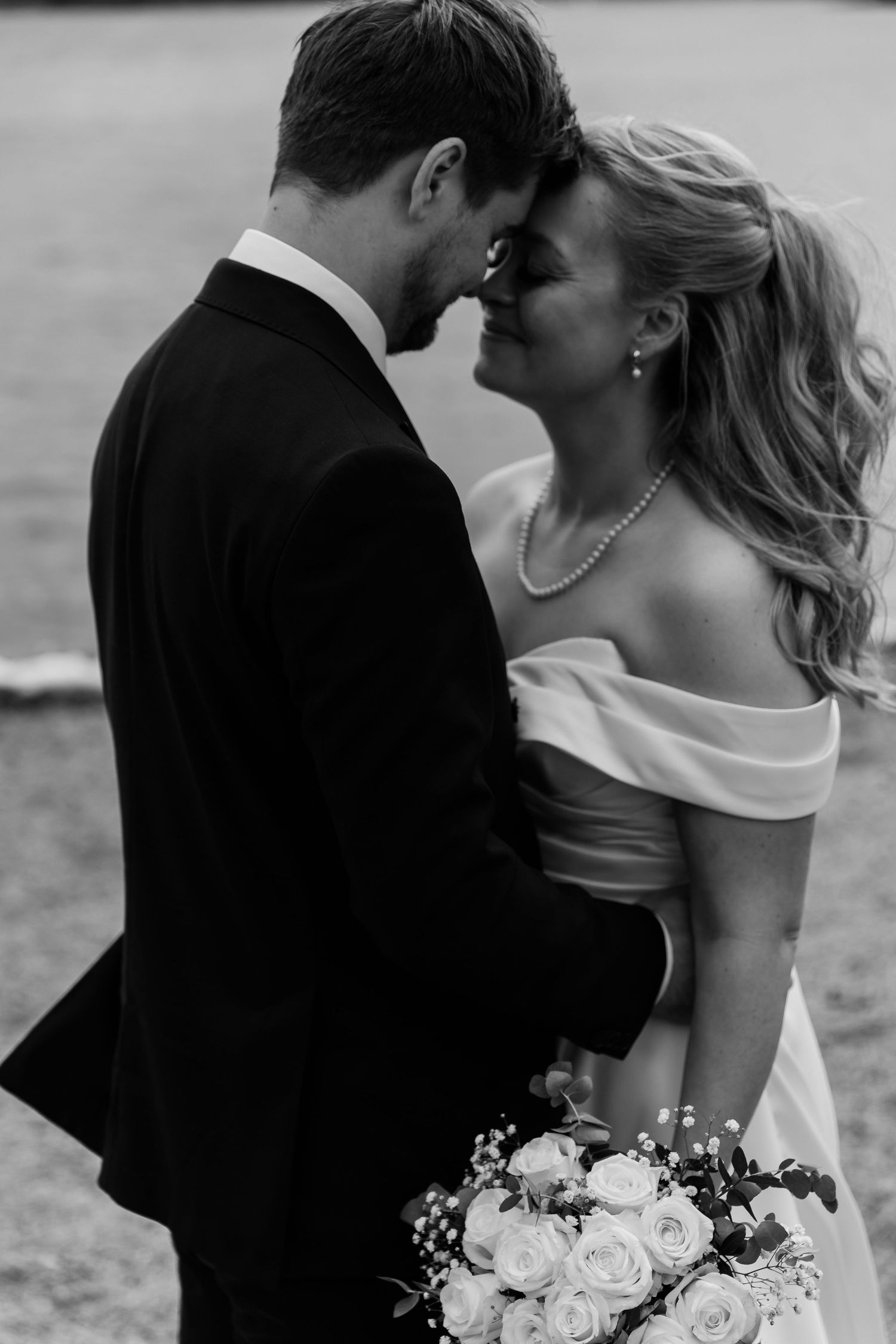 Black and white photo of bride and groom touching foreheads, bride holding white rose bouquet.