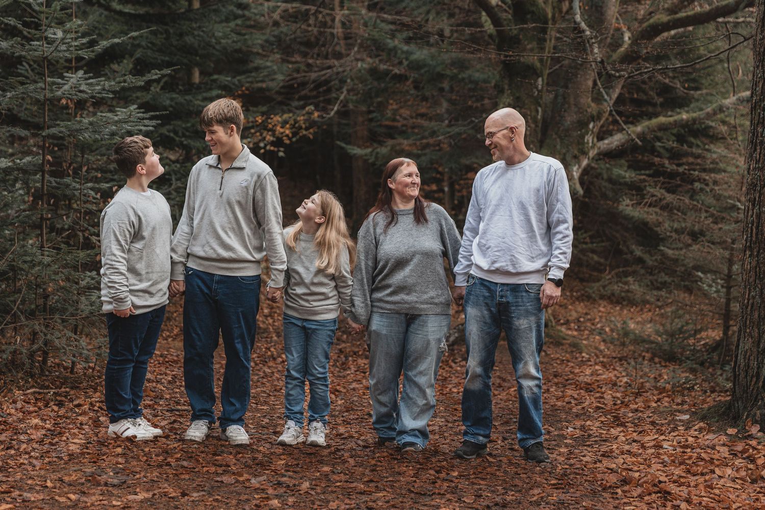 A family of five stands together on fallen autumn leaves in a wooded forest setting wearing casual grey sweaters.