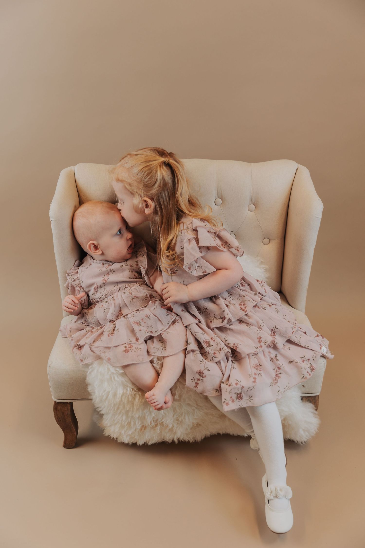 Two children in matching floral dresses sitting together on a beige tufted armchair against neutral background.