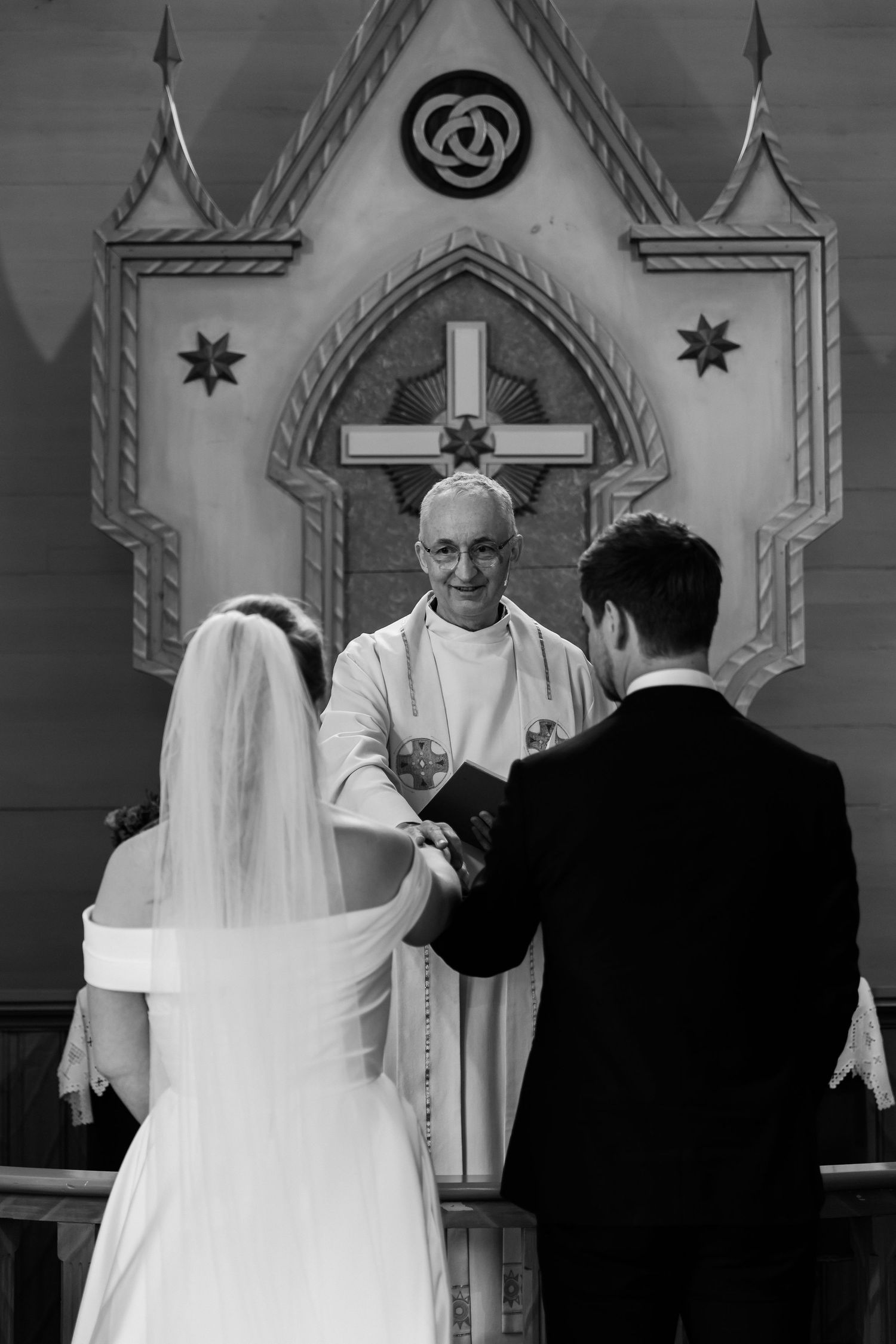 Black and white photo of a bride and groom exchanging vows before a smiling priest at a church altar.