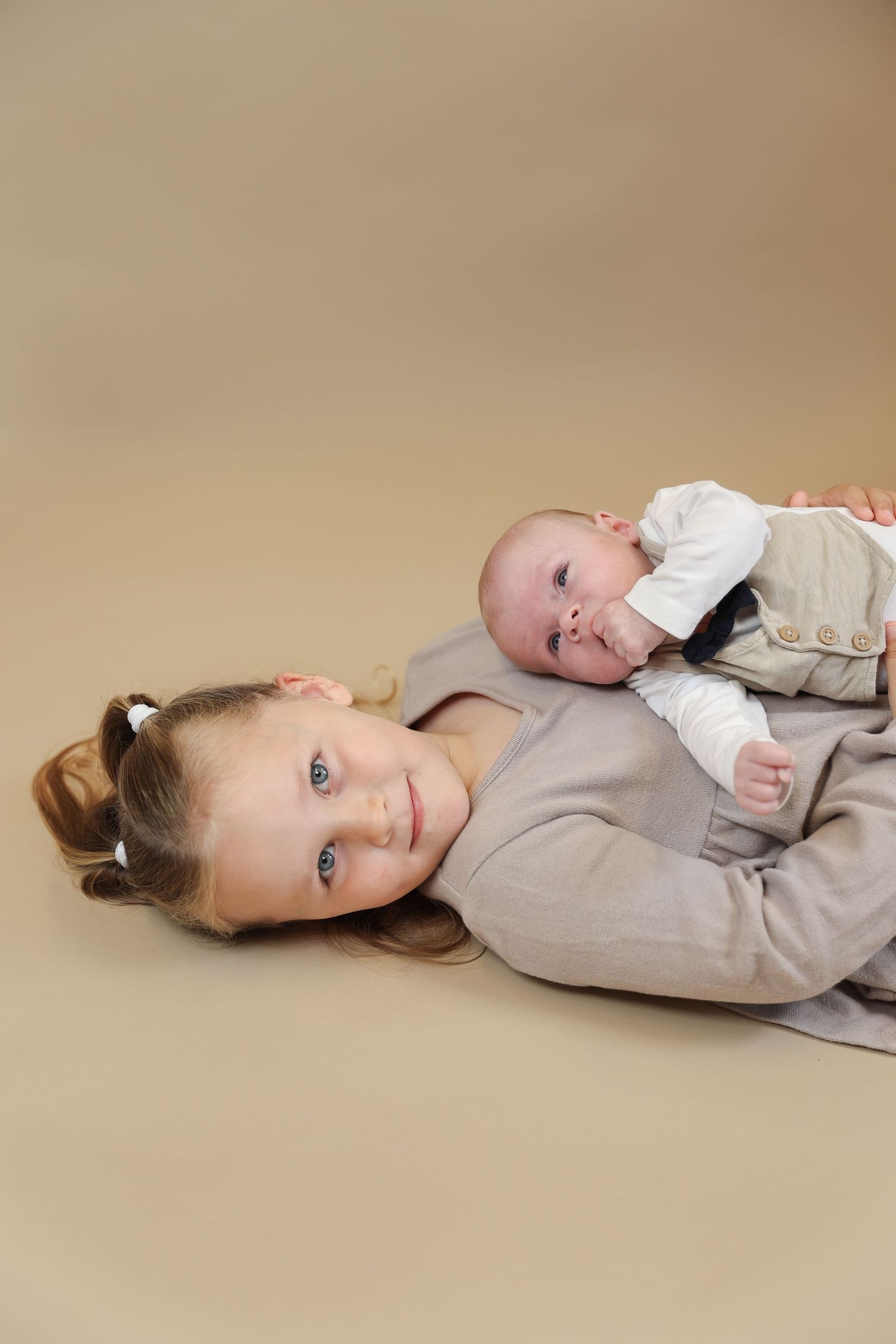 Two children lying together on beige background wearing light colored formal clothing.