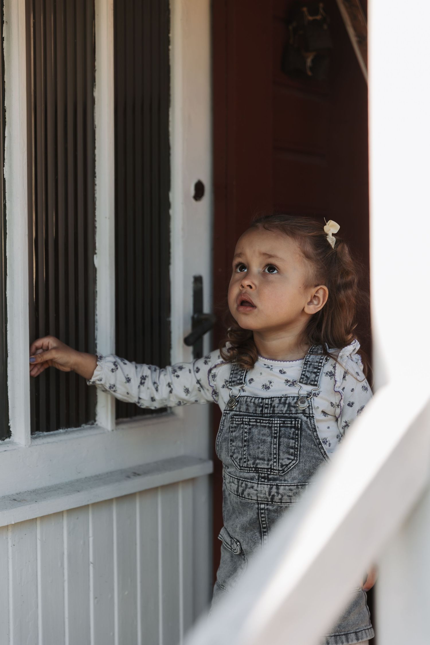 A small child in gray overalls peers out of a window with curiosity while standing indoors.