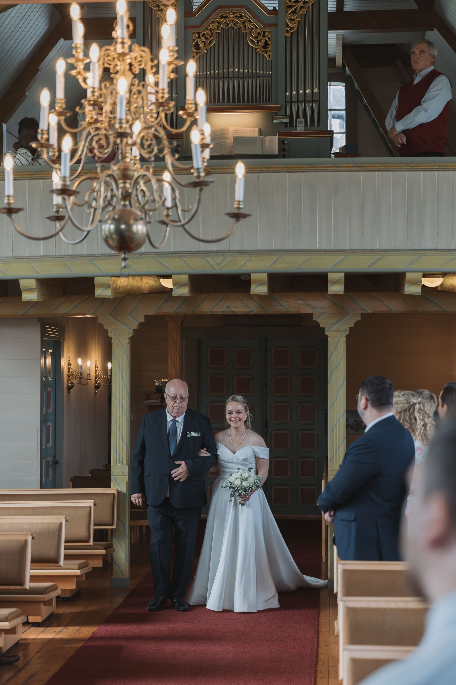 Bride walks down the aisle with escort in a charming wooden church with chandelier and pipe organ.