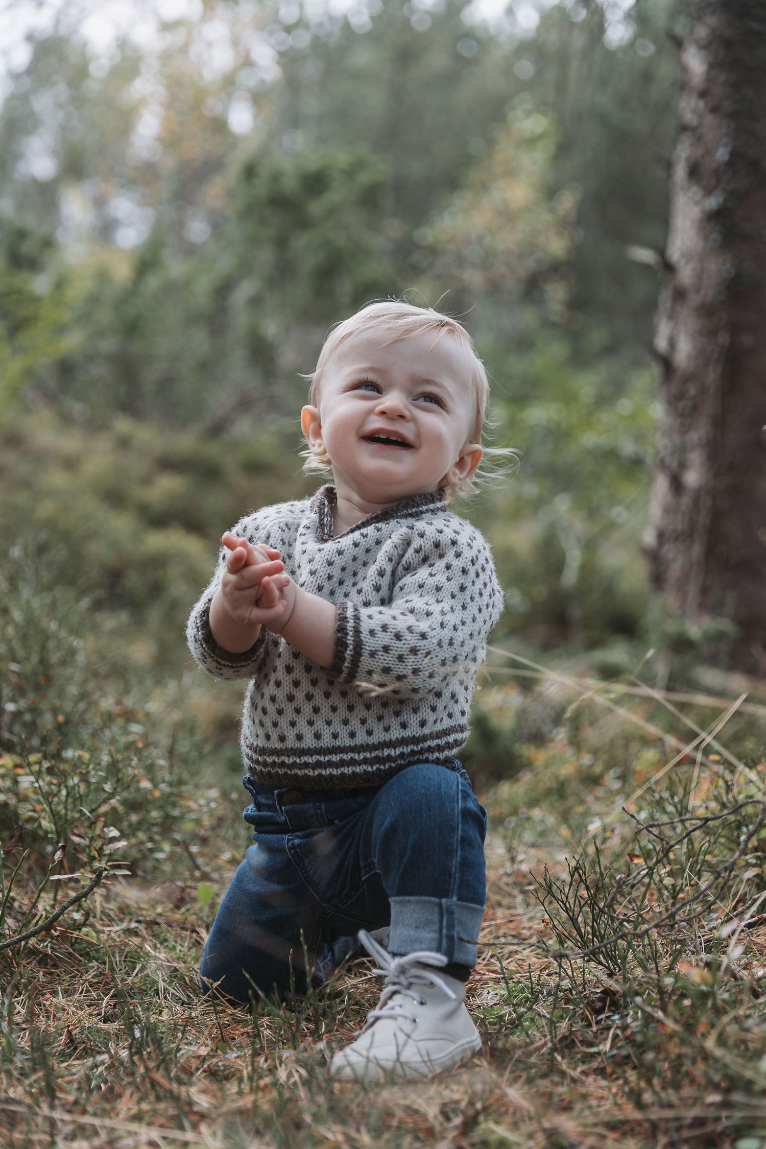 Happy toddler in gray sweater and jeans clapping hands outdoors in a forest setting.