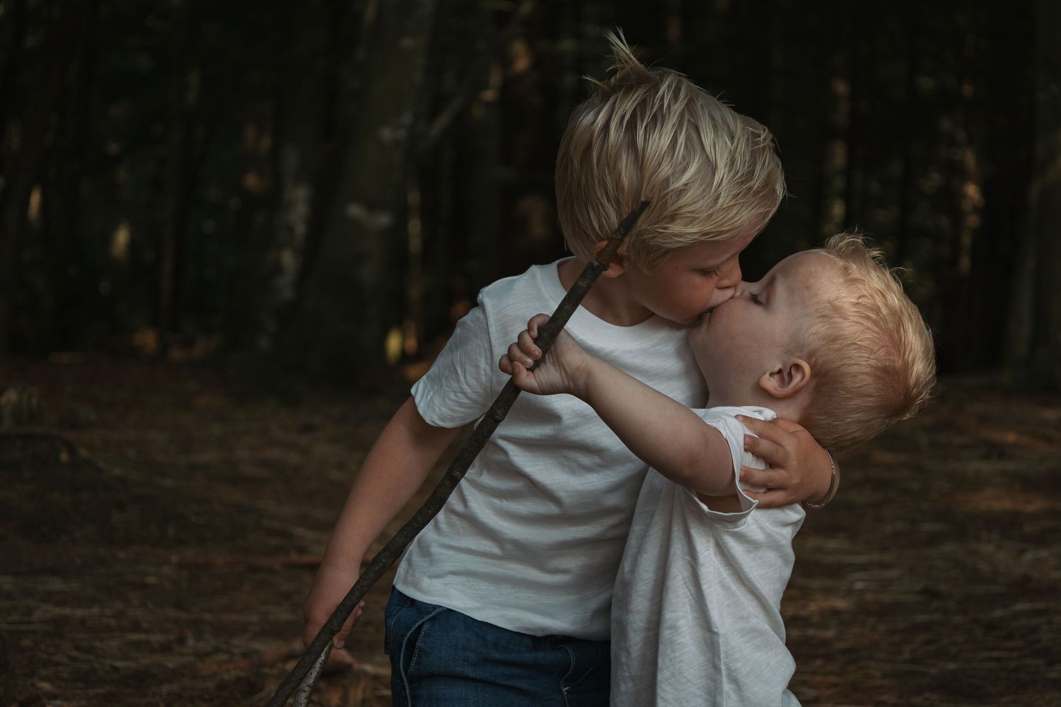 Two young blonde siblings sharing a tender kiss in a forest setting, wearing white t-shirts.