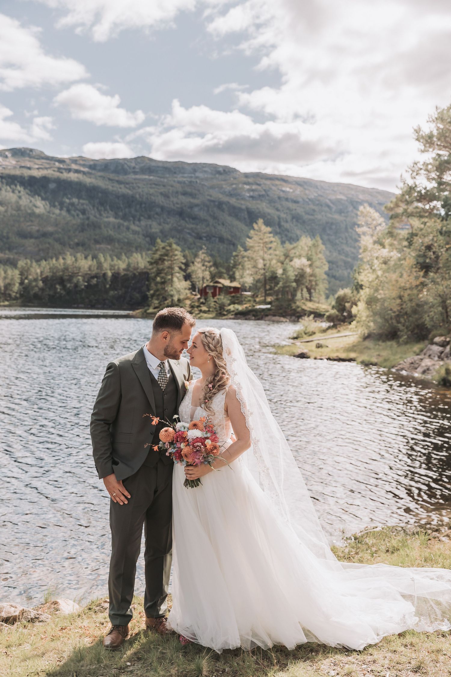 Bride and groom share a romantic moment by a scenic mountain lake, bride holding a colorful bouquet in a white gown.