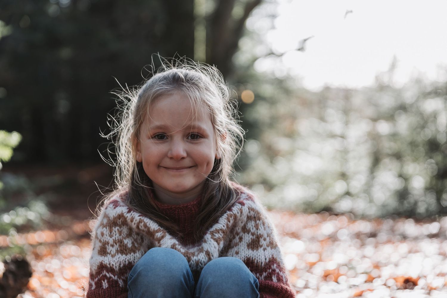 A child wearing a cozy patterned sweater sits among autumn leaves on a sunny fall day.