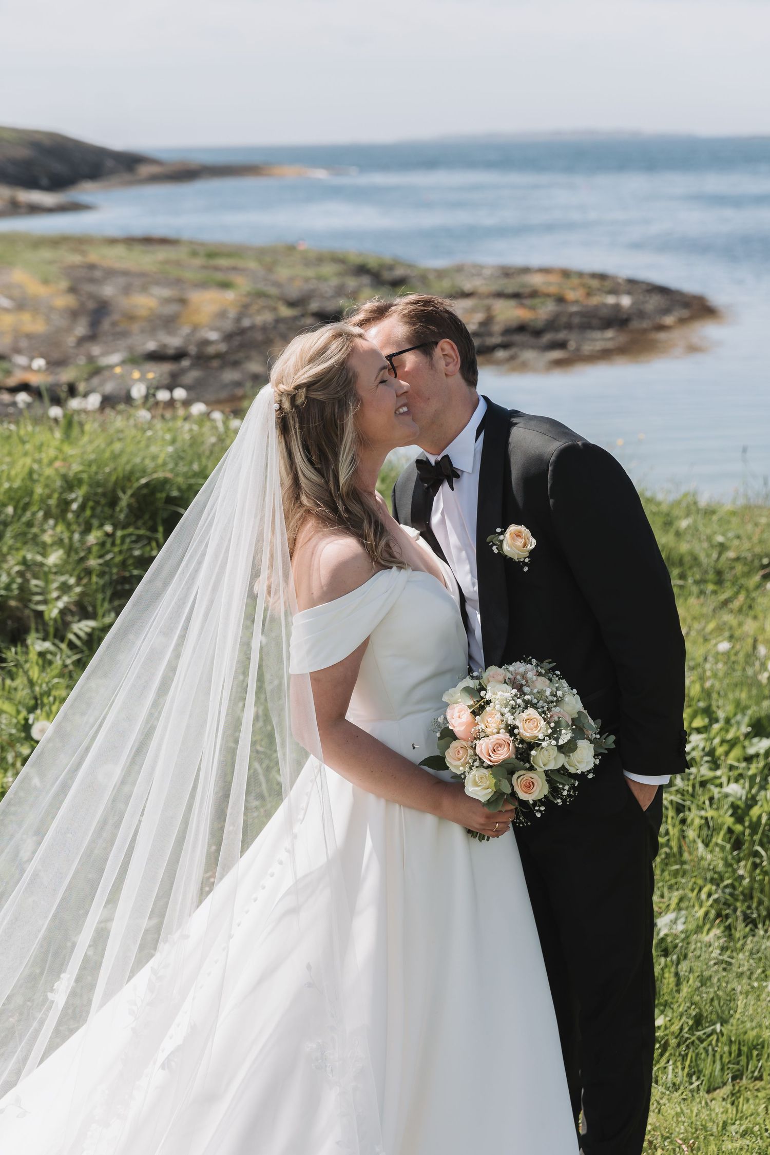 Bride and groom kissing outdoors near rocky coastline, bride in off-shoulder gown with veil holding pastel bouquet.
