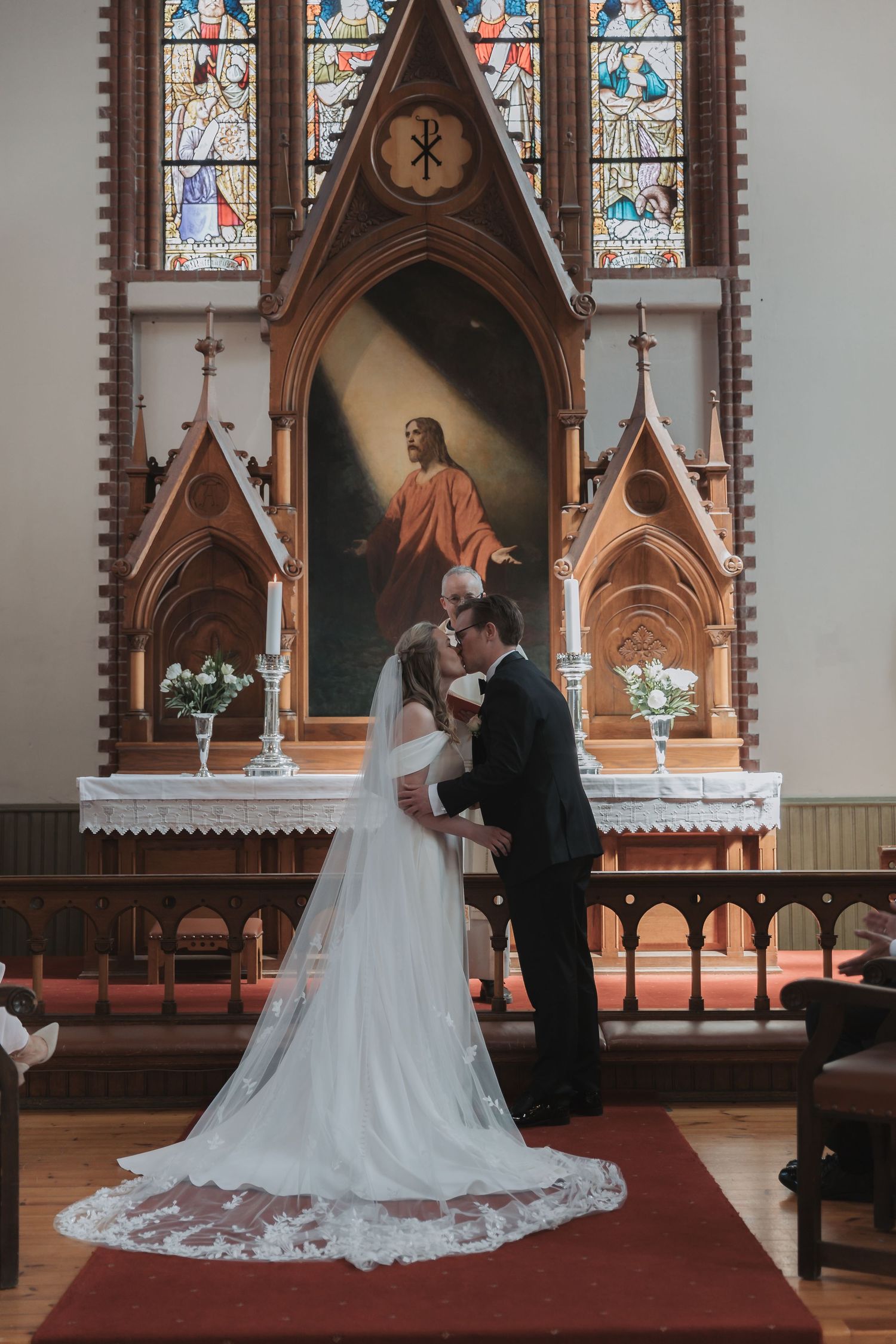 Bride and groom share first kiss at church altar with stained glass windows and Jesus painting backdrop.