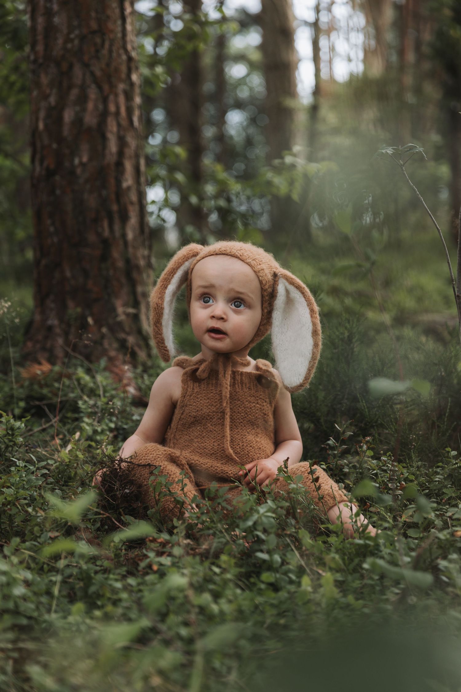 Adorable baby in brown bunny costume with floppy ears sitting in a lush green forest setting.