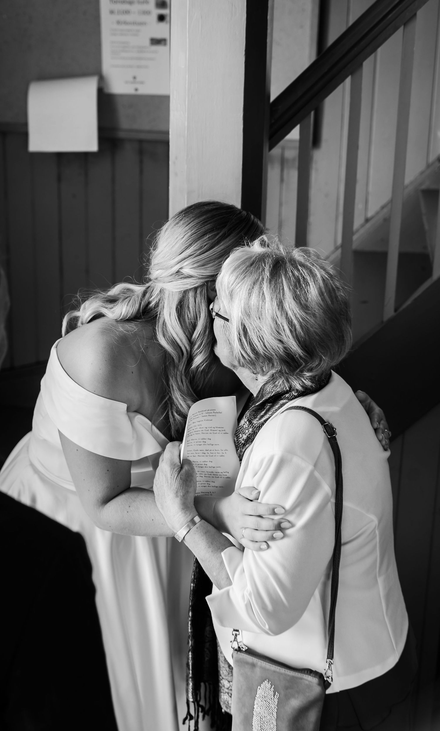 Black and white photo of a bride in white dress sharing an emotional hug with an older woman at a wedding.