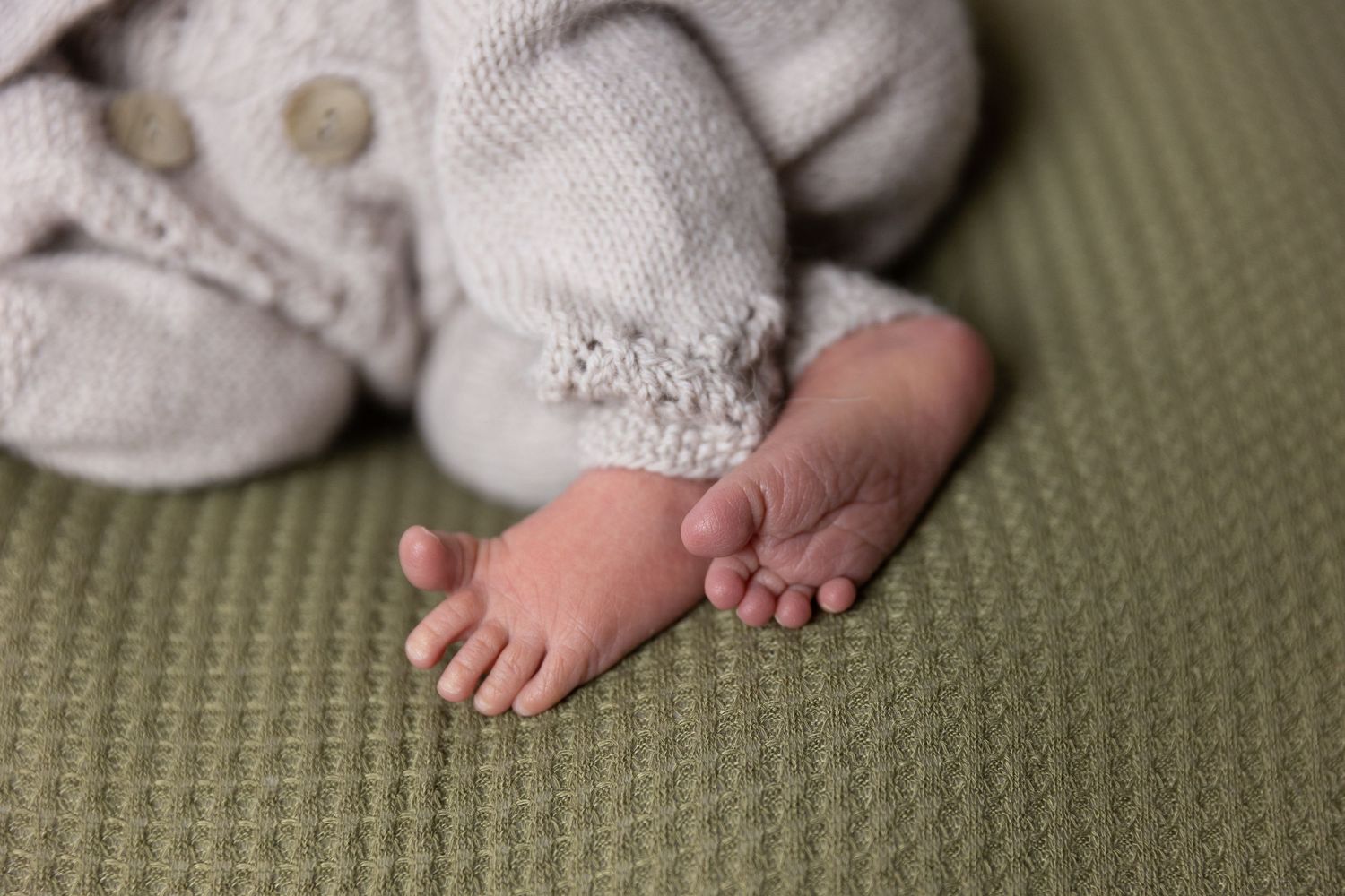 Tiny bare baby feet peeking out from white knitted clothing on green textured background.