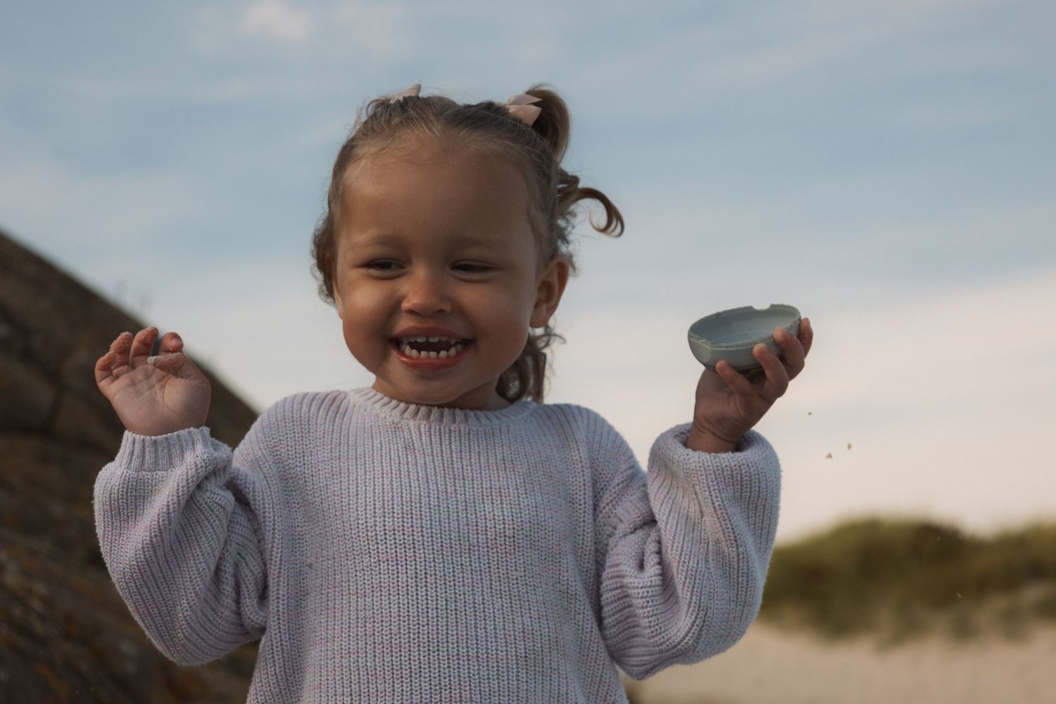 Joyful child in light sweater holds something while smiling against cloudy sky.