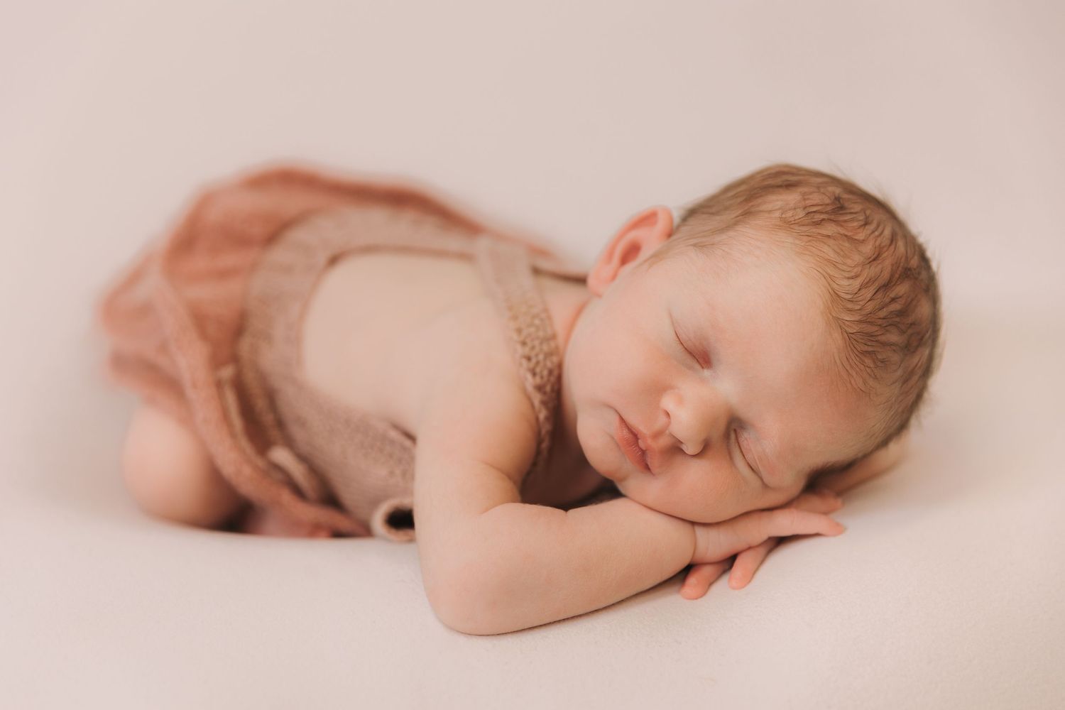 Peaceful sleeping baby in soft pink tones lying on textured blanket in artistic newborn photography series.
