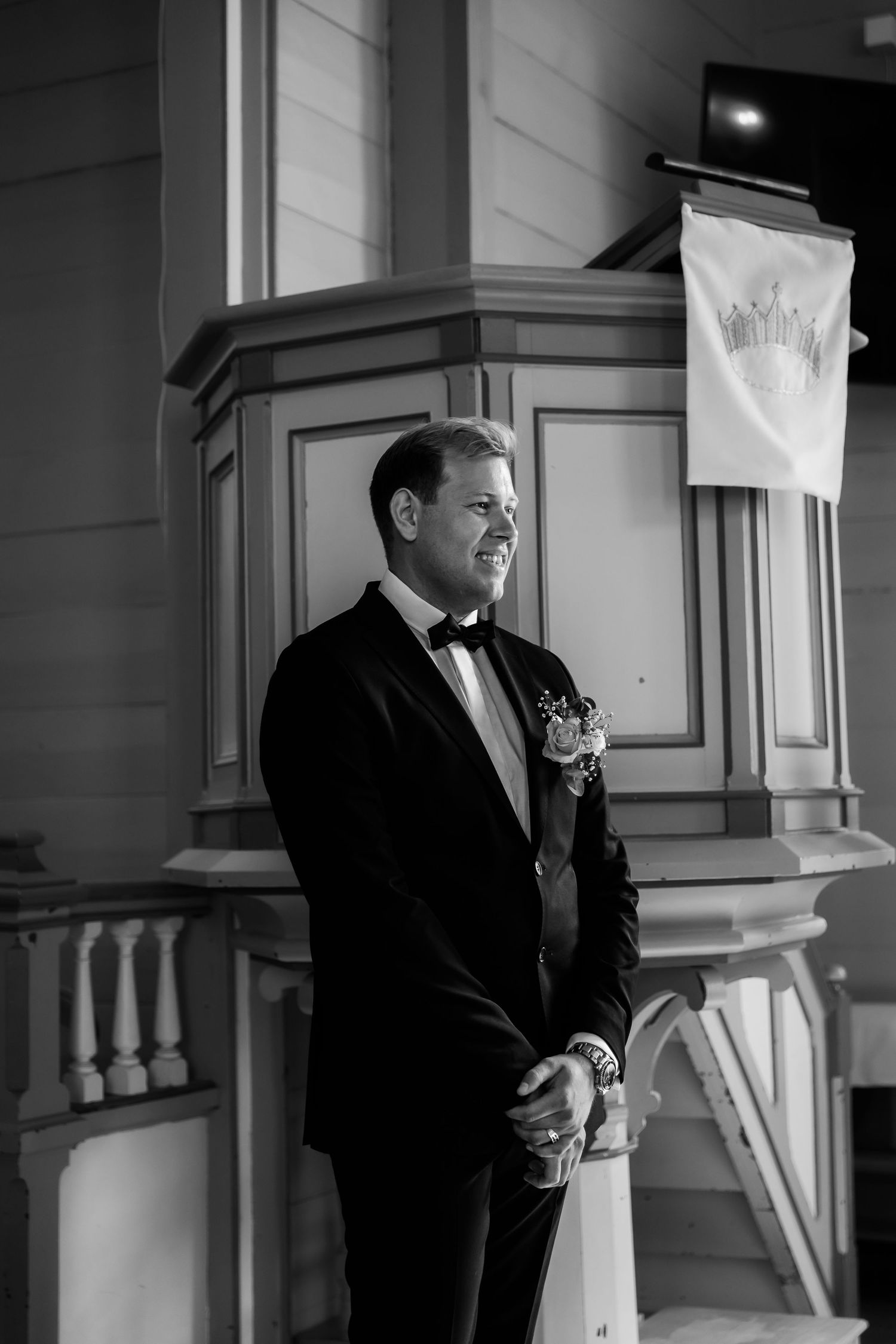 Black and white photo of a groom in a tuxedo with boutonniere standing at the altar in a church.