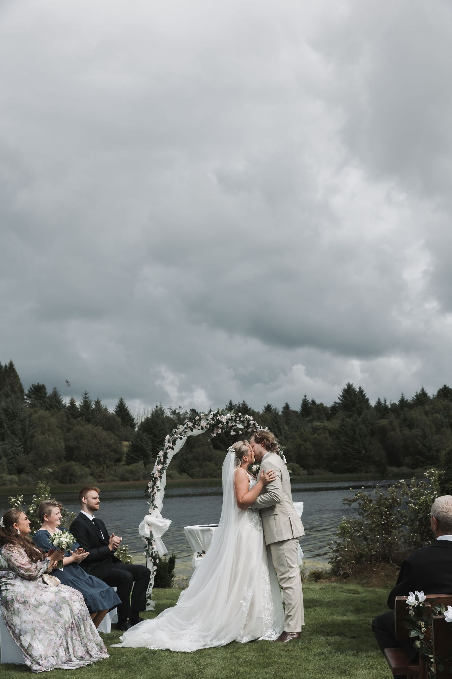 Couple shares first kiss at outdoor wedding ceremony by a lake, framed by a floral arch with guests seated nearby.