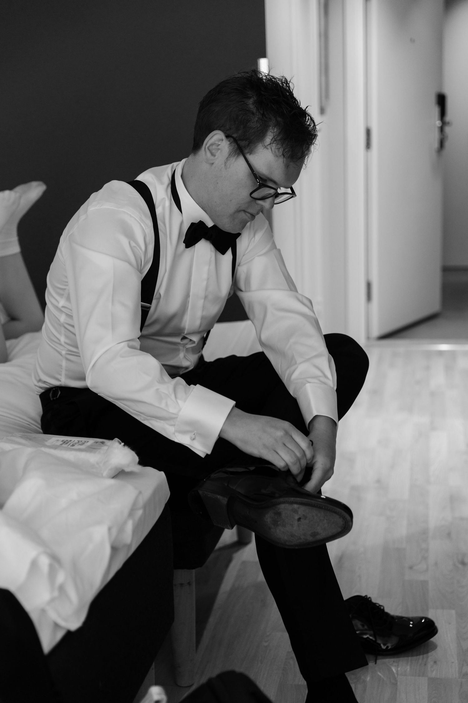 Groom in white shirt and bow tie putting on dress shoes while sitting on bed before wedding ceremony.