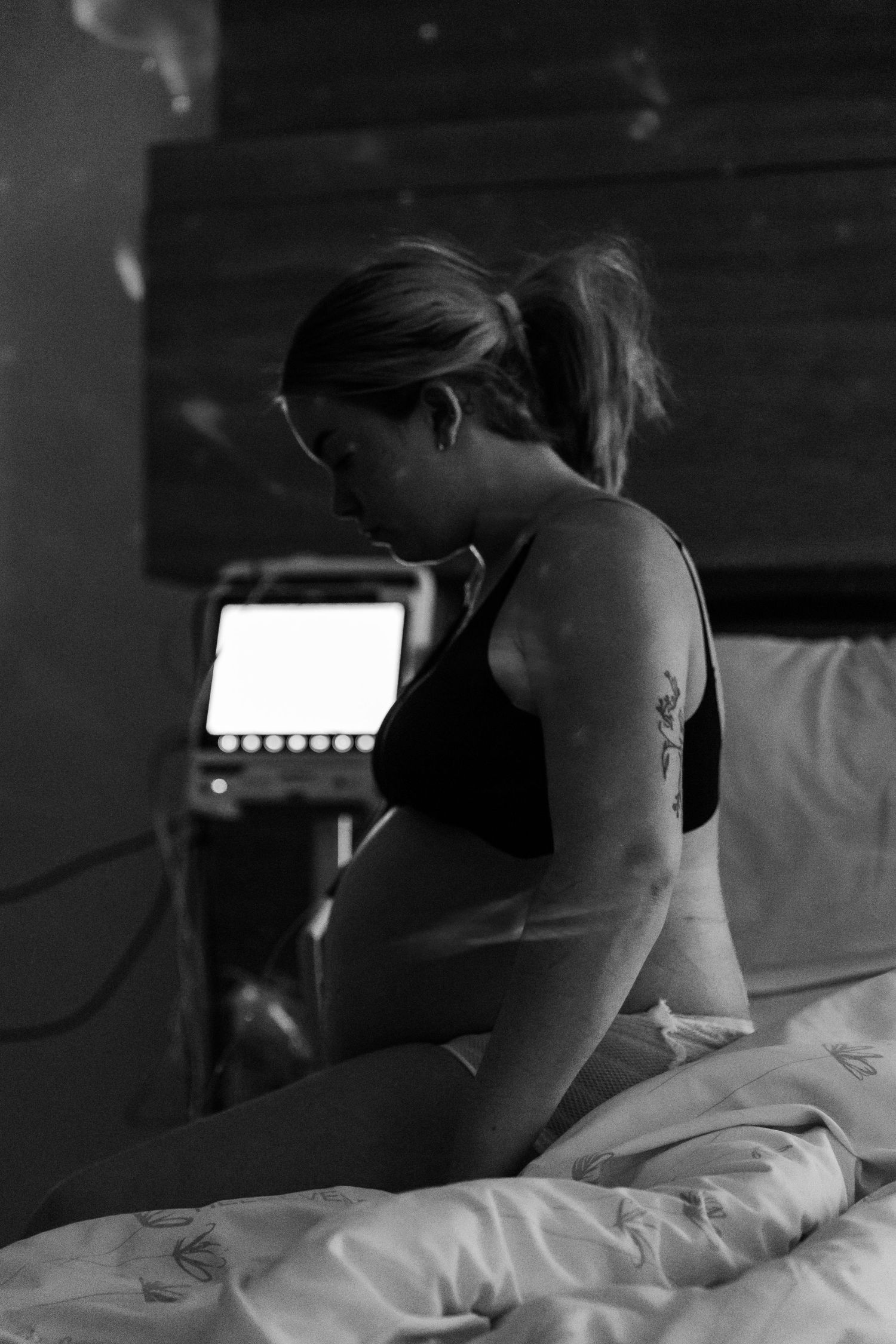A person sits on a bed in a dark room illuminated by the glow of a laptop screen in black and white.