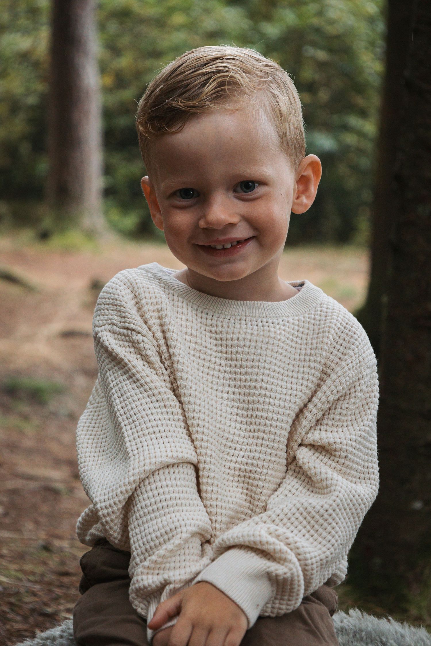Young child in cream knit sweater smiles while sitting outdoors in a forest setting.