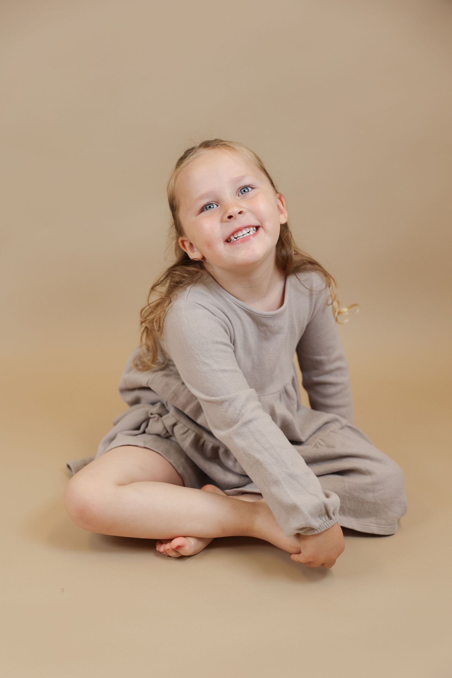 A smiling child in a gray dress sits cross-legged on a beige backdrop during a portrait photography session.