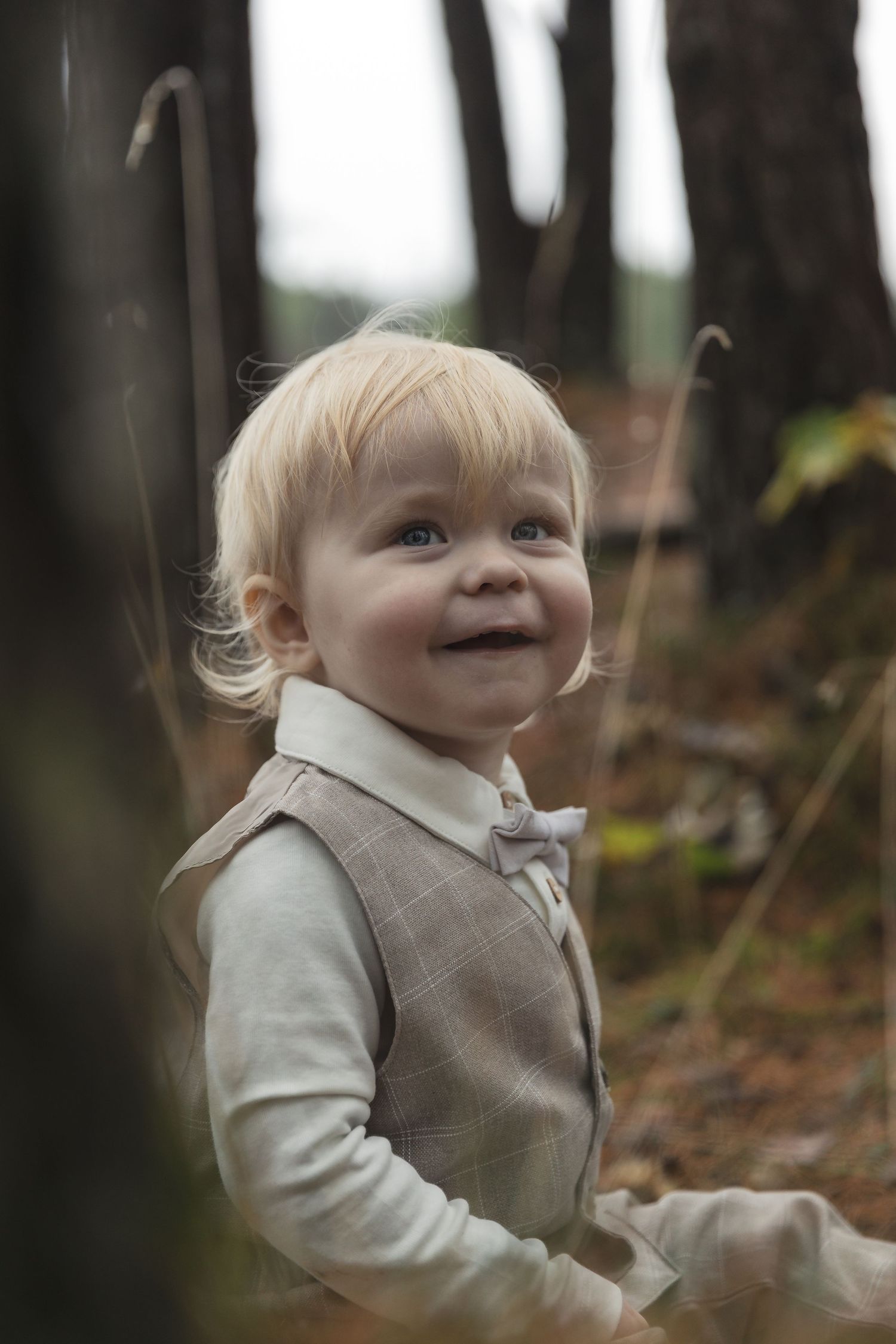 Smiling blonde toddler boy wearing a plaid vest and bow tie in a forest setting.