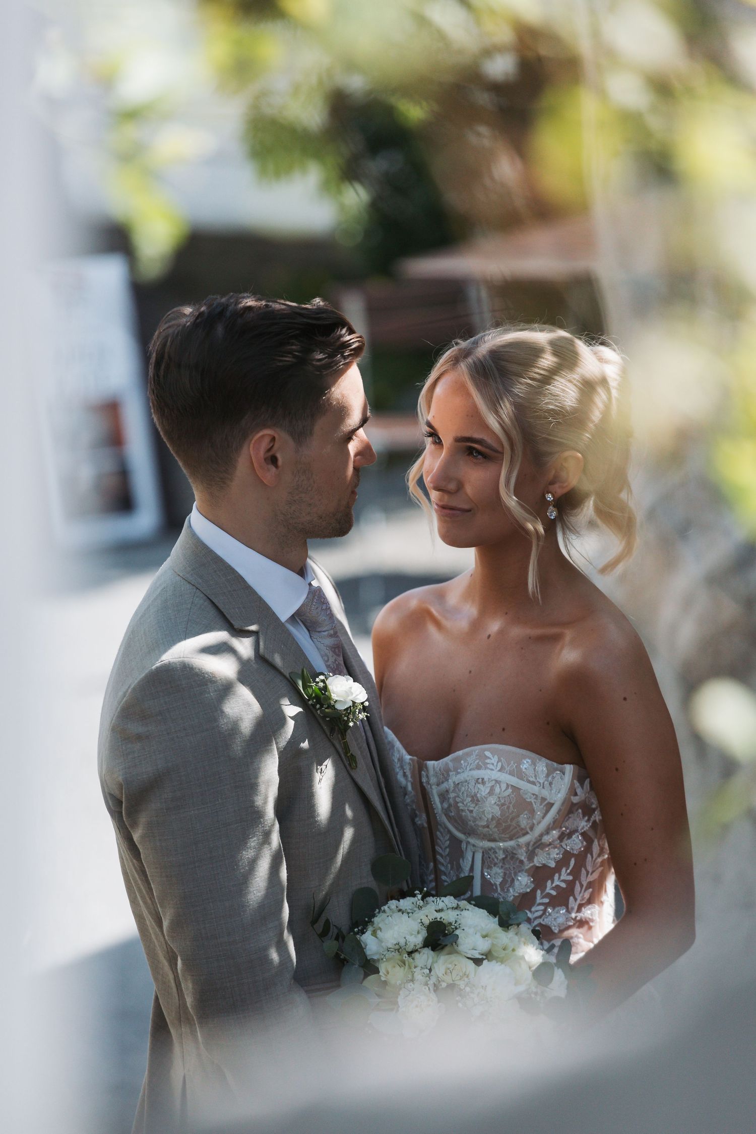Bride and groom share an intimate moment, she holds white bouquet, he wears grey suit with boutonniere.
