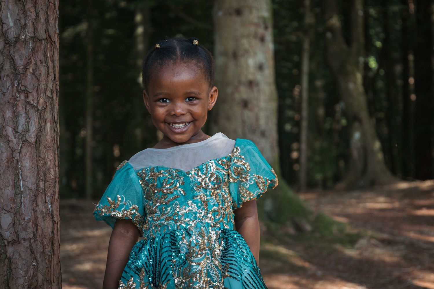 Cheerful child wearing turquoise dress with intricate patterns stands in wooded setting.