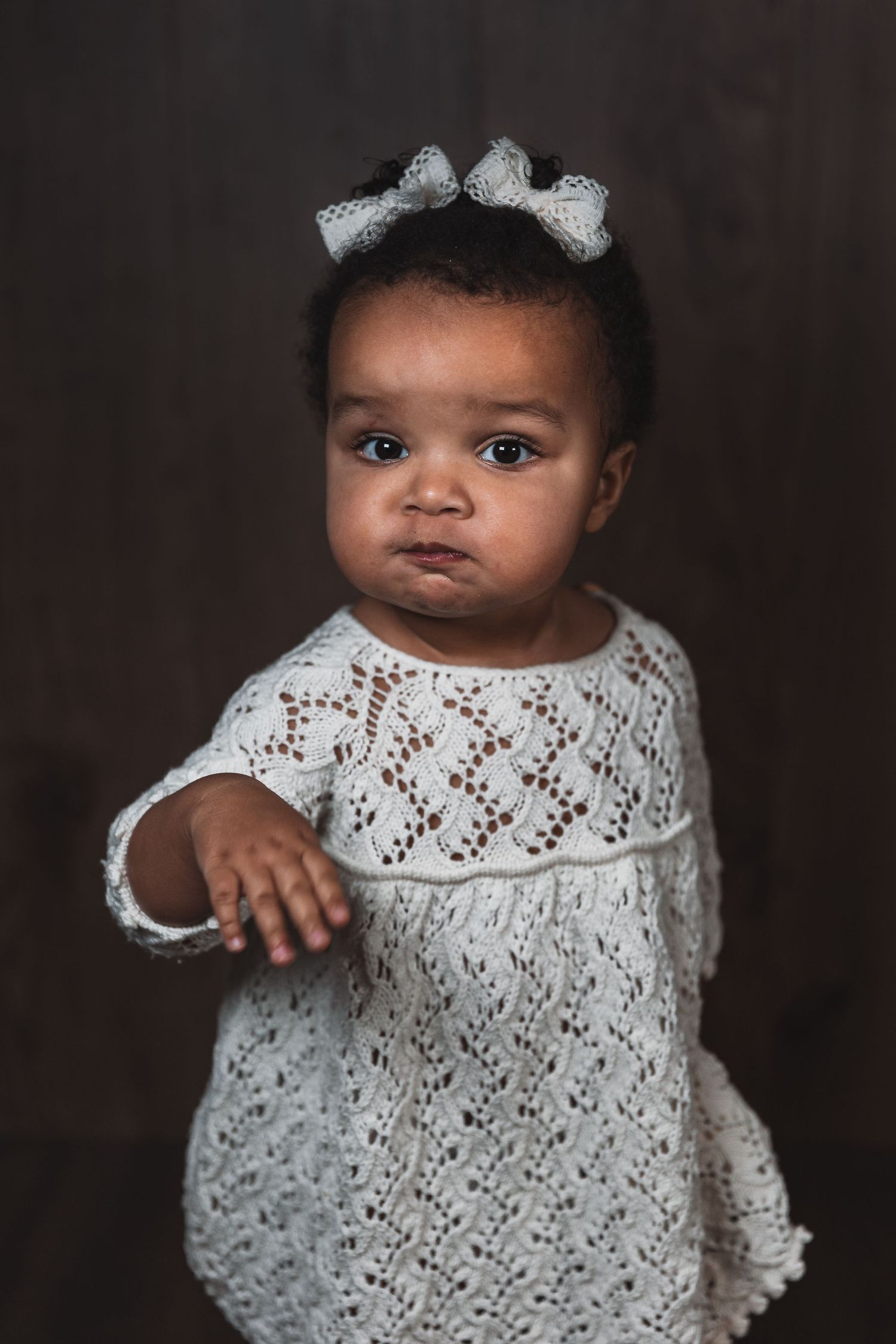 Baby wearing white lace dress with bows in hair against dark background for portrait session.