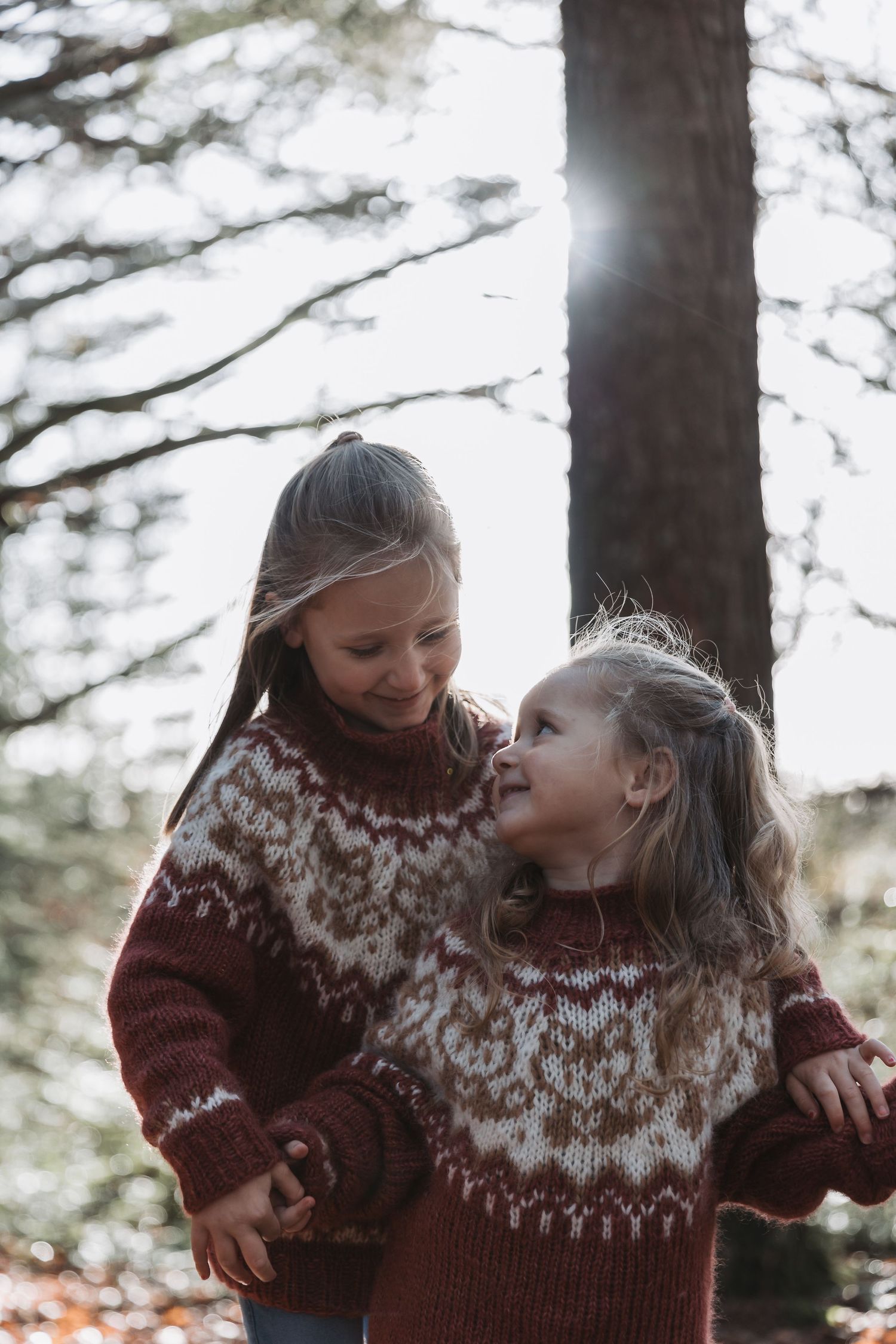 Two girls in matching Nordic sweaters smile together in a sunlit forest setting.