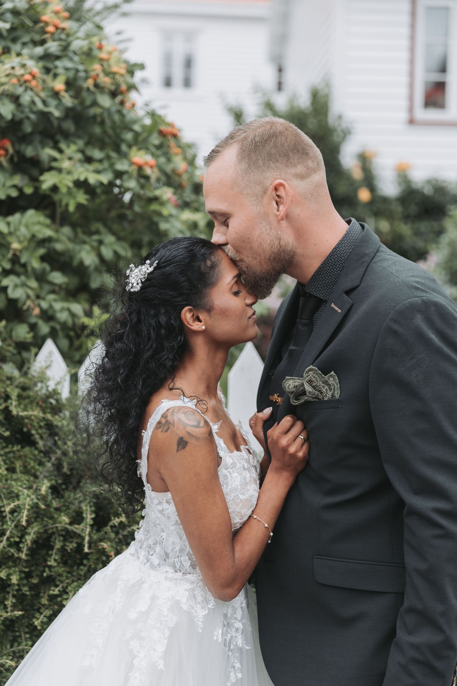 Interracial couple sharing a romantic kiss in wedding attire, bride in lace gown with groom in dark suit outdoors.