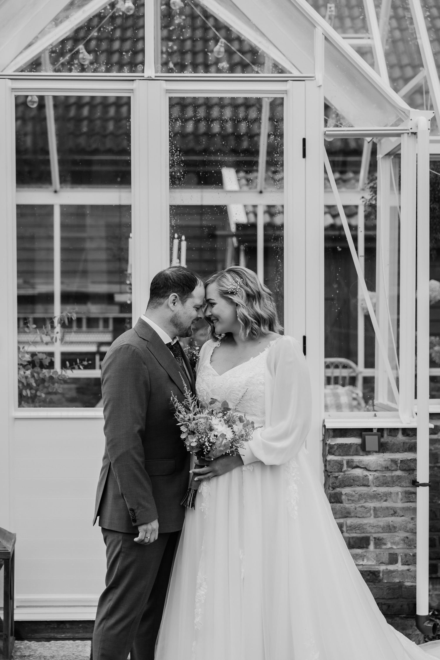 Black and white photo of a bride and groom sharing a romantic moment in front of a greenhouse on their wedding day.