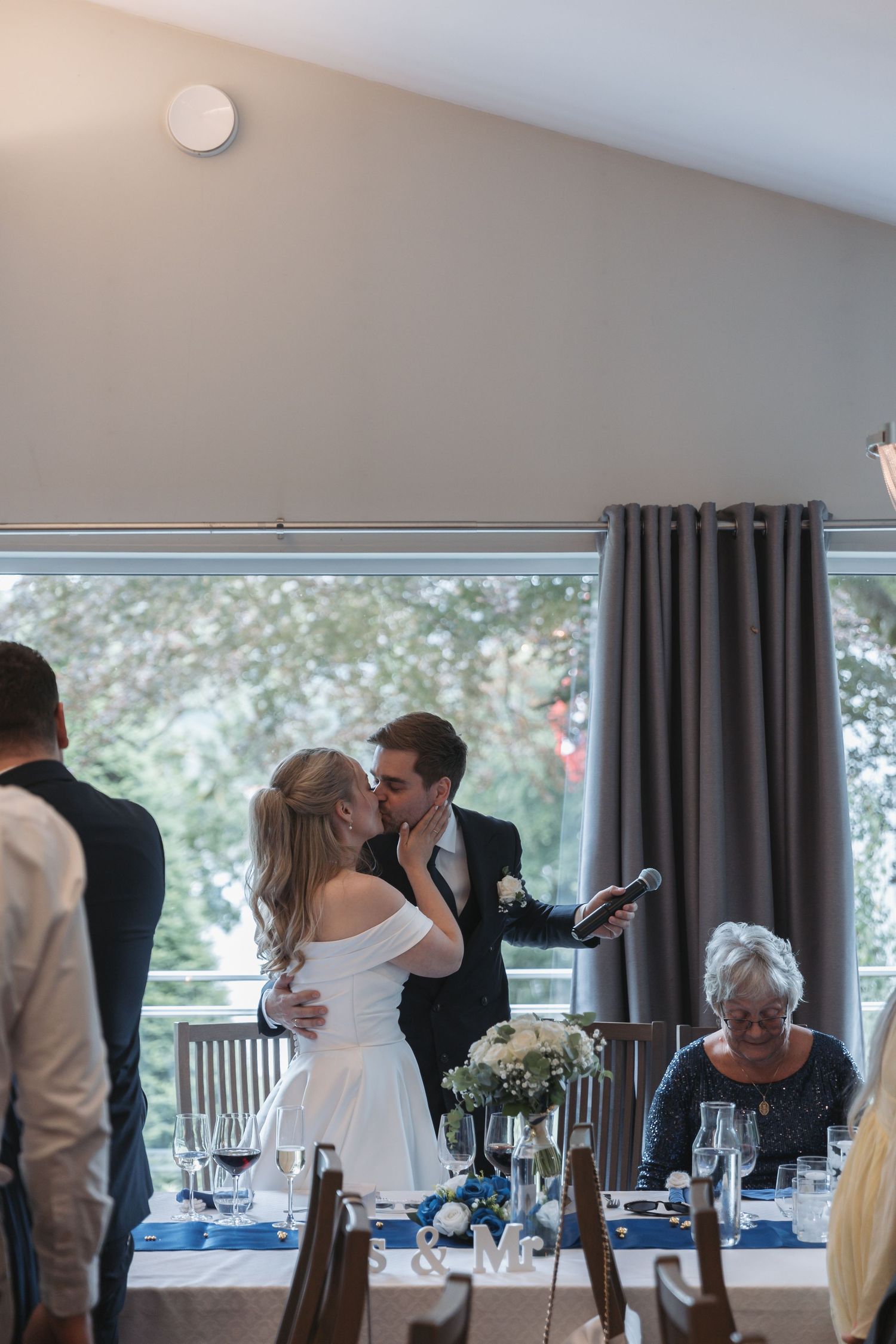 Bride and groom share a kiss at their wedding reception, with guests seated at a decorated table with blue accents.