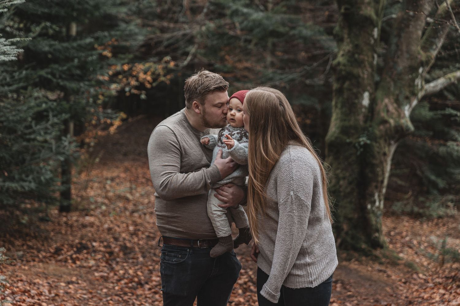 A couple in cozy sweaters holding a small baby while standing in an autumn forest with fallen leaves.
