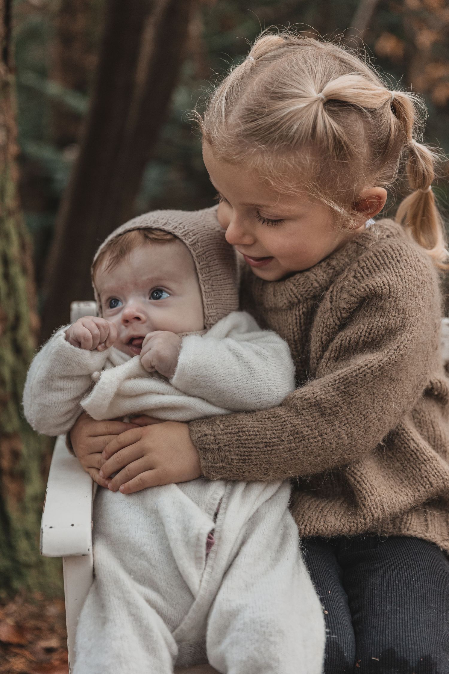 A child in a brown sweater cradles and interacts with a baby wearing a white outfit in a forest setting.