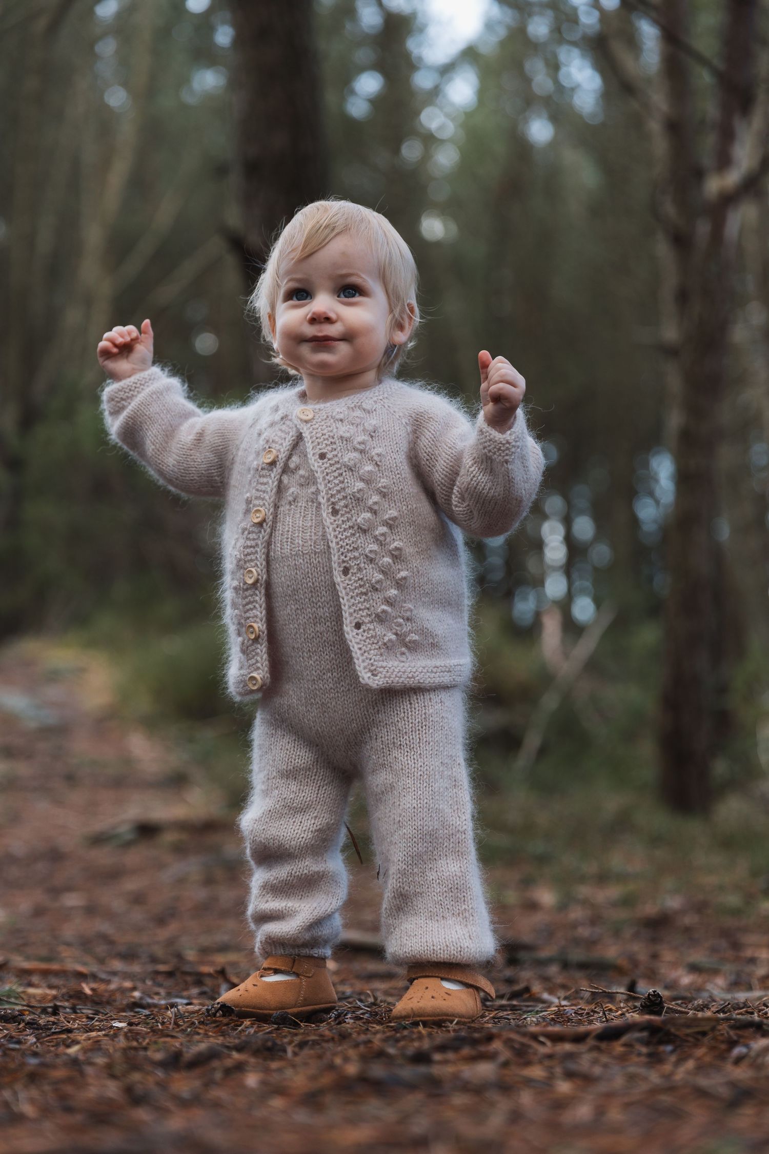 Small child wearing cozy knit beige outfit and moccasin shoes stands with raised arms on forest path.