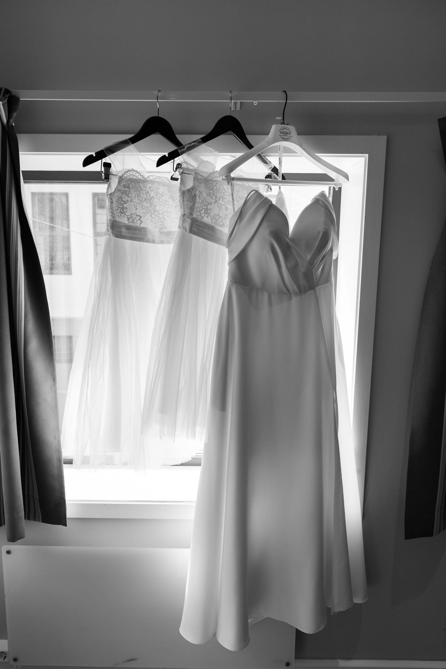 Black and white photo of three elegant wedding dresses hanging on hangers in front of a bright window.