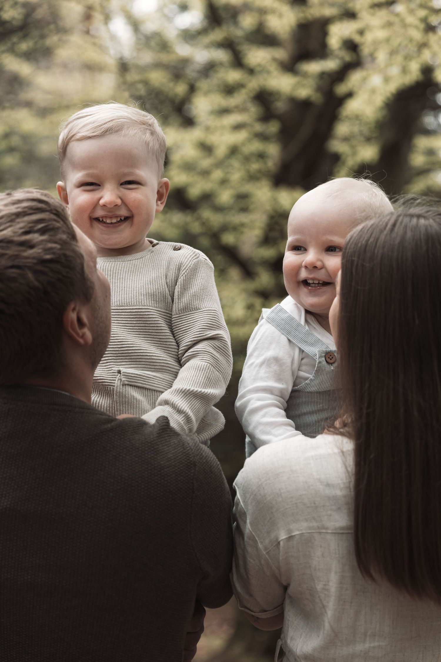 Two laughing toddlers held by their parents outdoors in a lush green park setting.