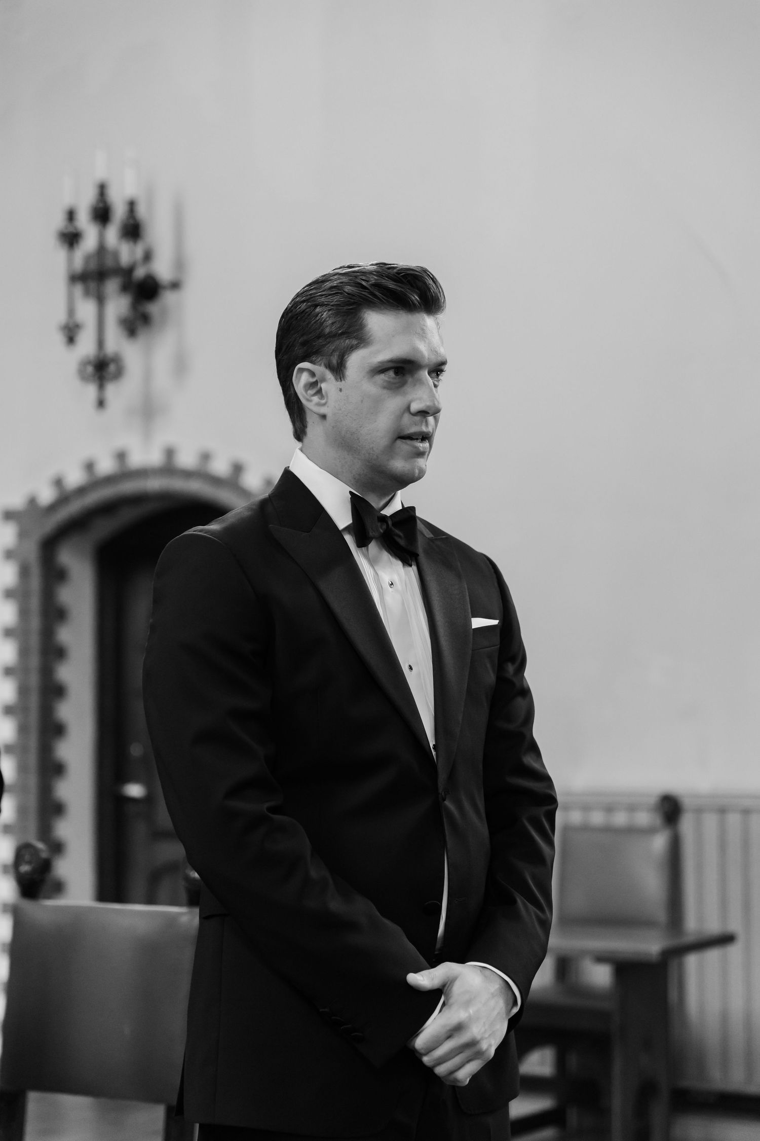 Elegant groom in black tuxedo and bow tie standing in a church, black and white wedding photography.