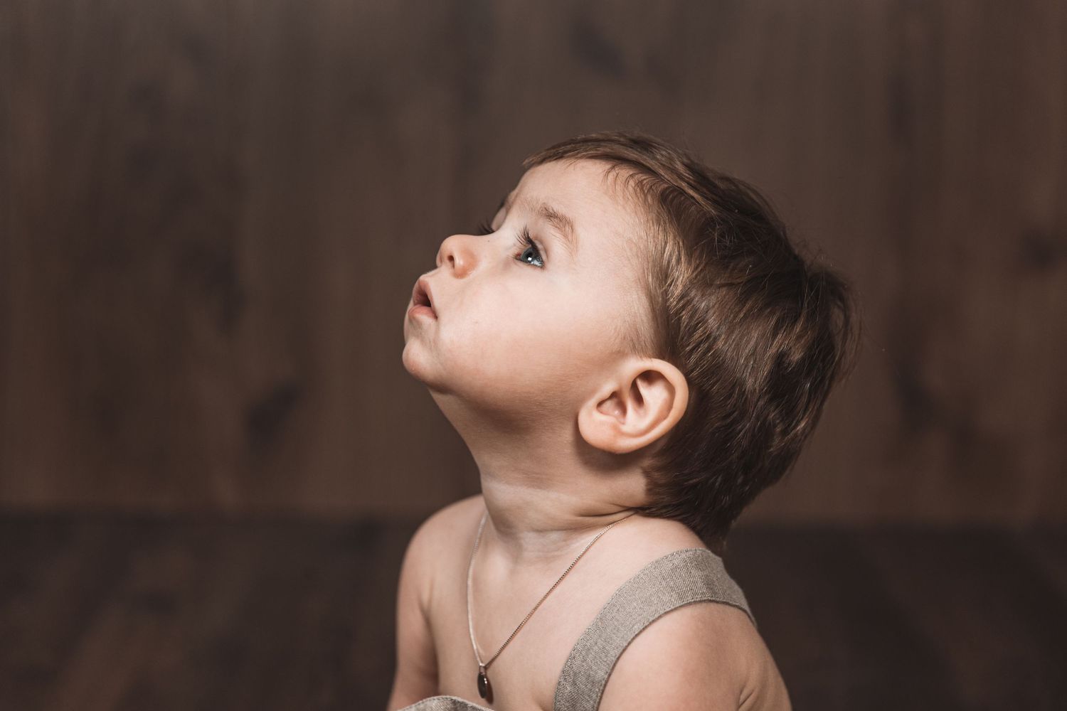 Small child in gray outfit looks upward with curiosity against dark background.