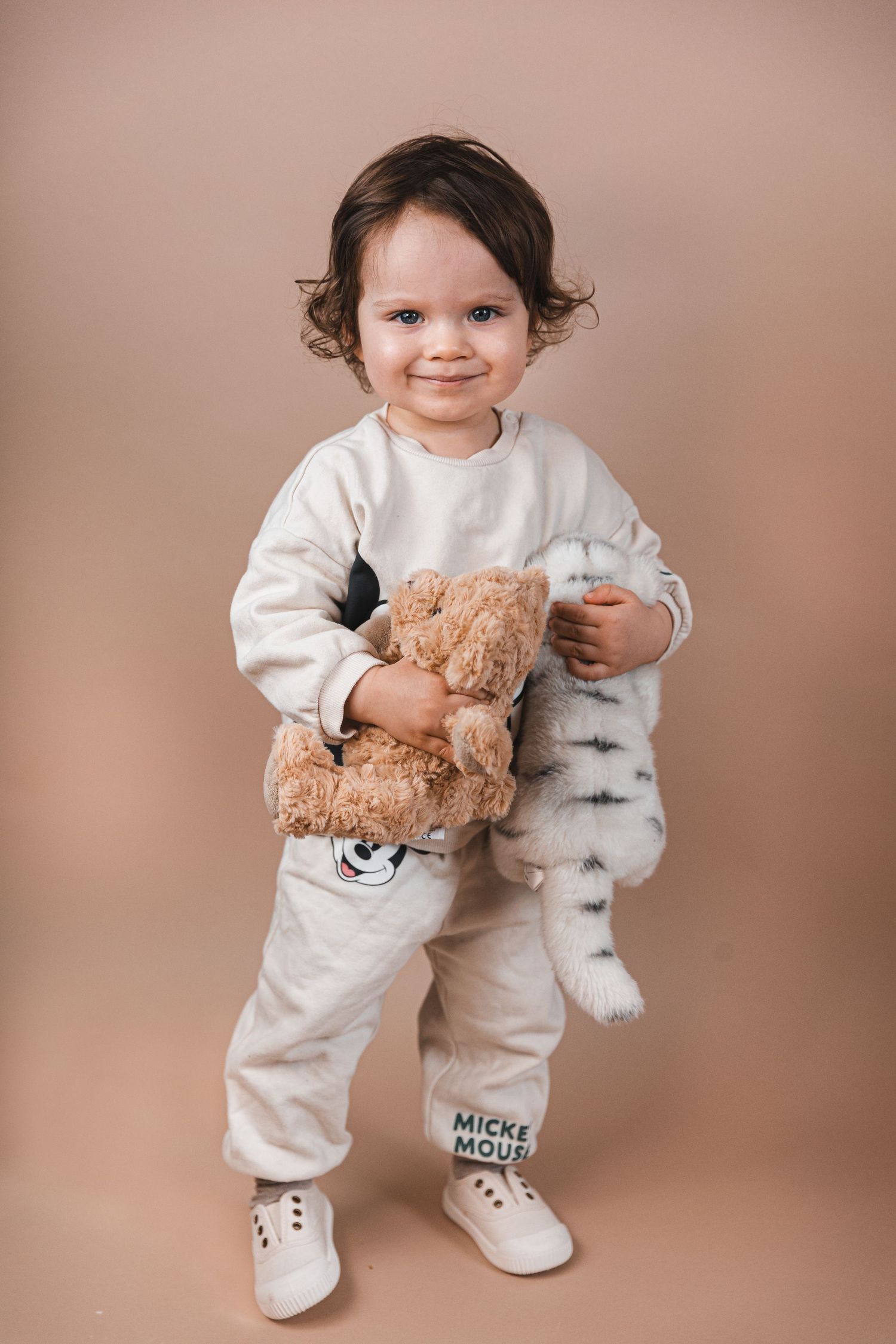 A small child in a cozy white outfit holds a teddy bear while standing against a neutral brown background.