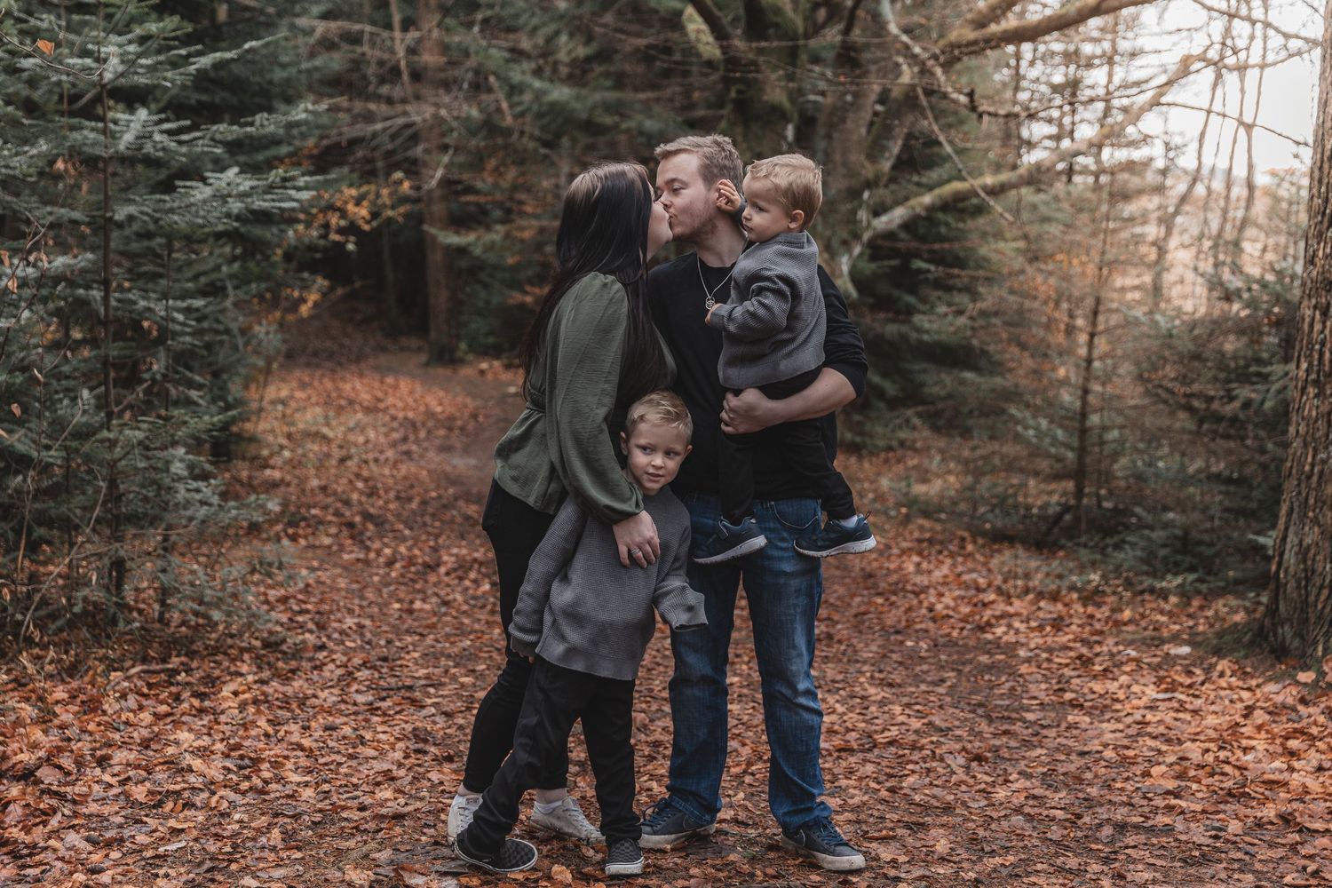 A family of four stands together on an autumn trail surrounded by evergreen trees and fallen orange leaves.