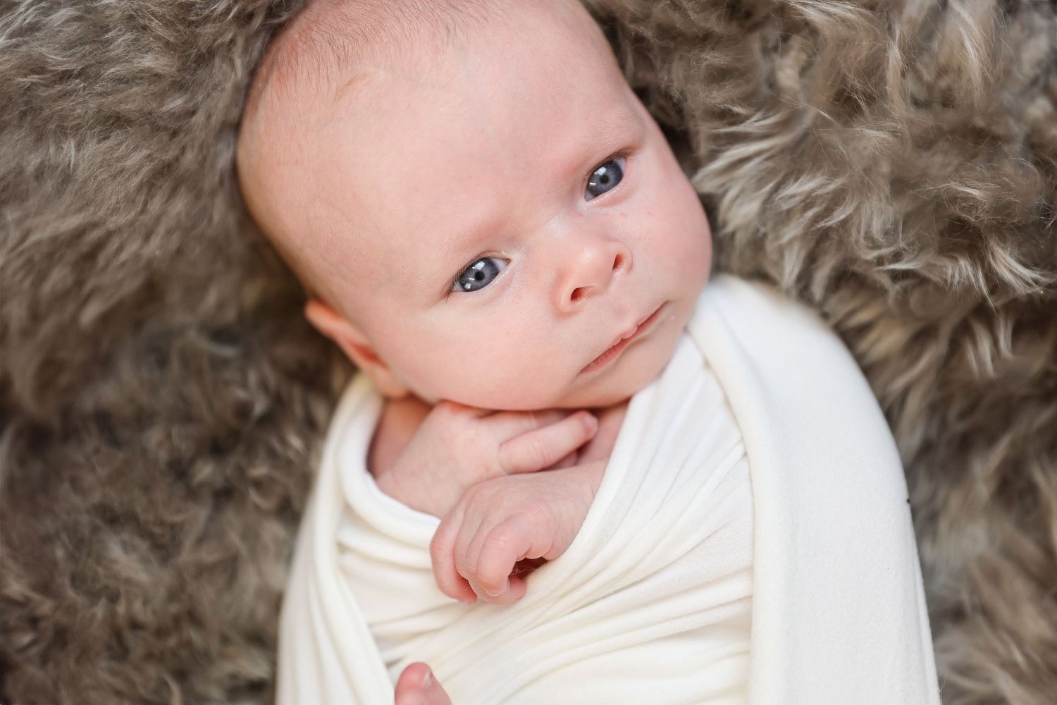 Baby wearing white outfit smiles while lying on soft gray fur blanket.