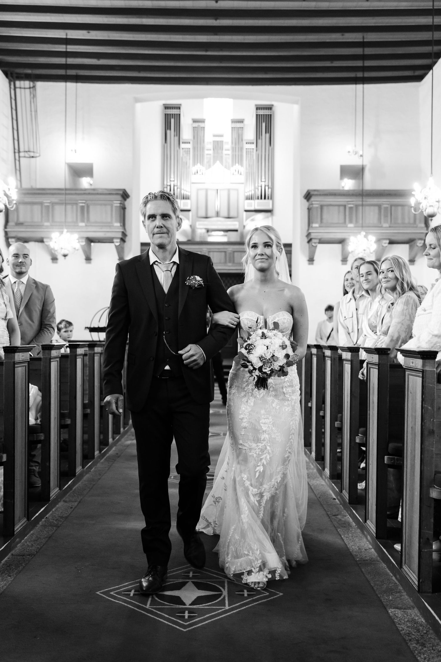 Black and white photo of a bride in lace gown walking down church aisle with escort during wedding ceremony.