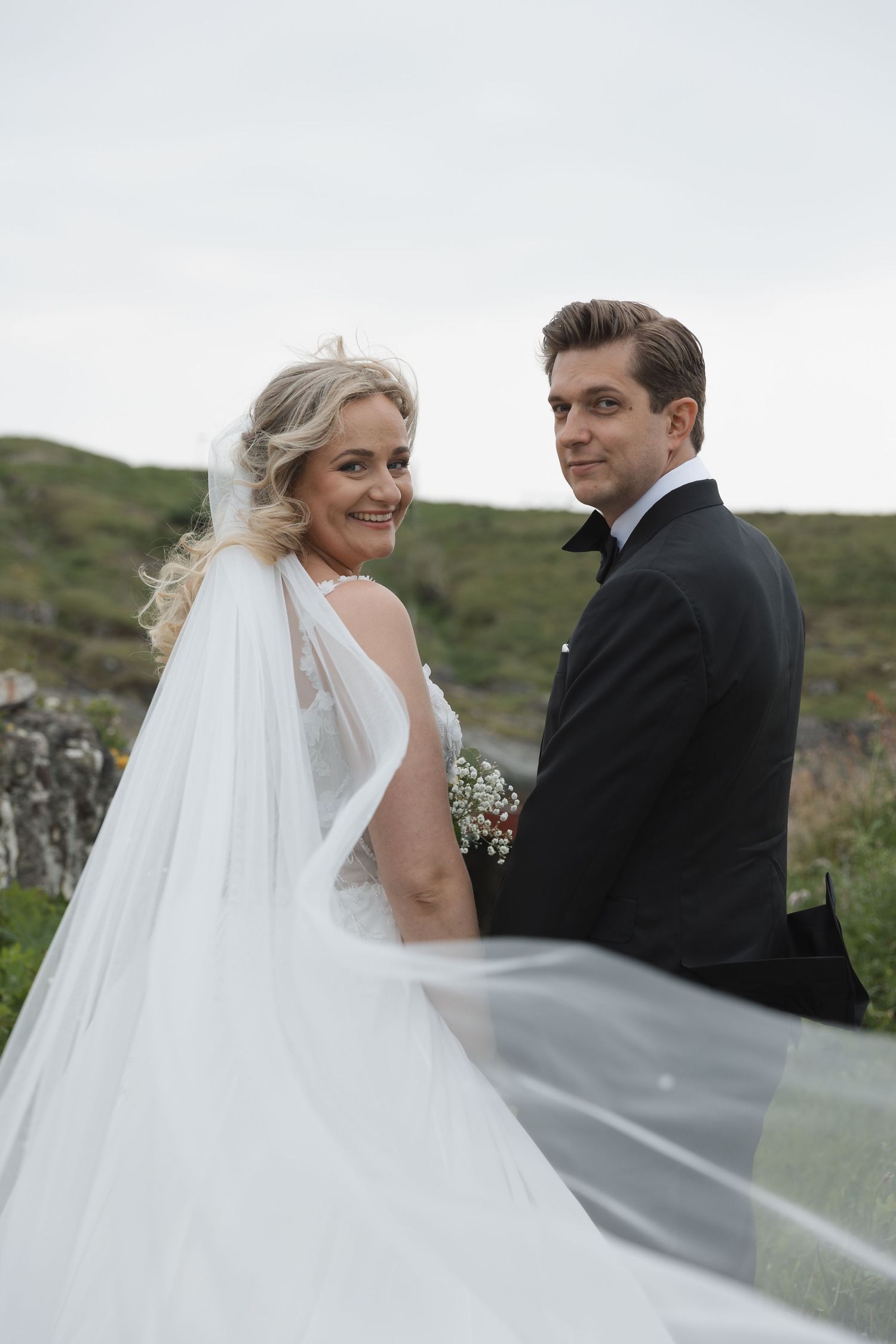 A bride in a flowing white gown and veil poses with a groom in a black tuxedo outdoors on their wedding day.