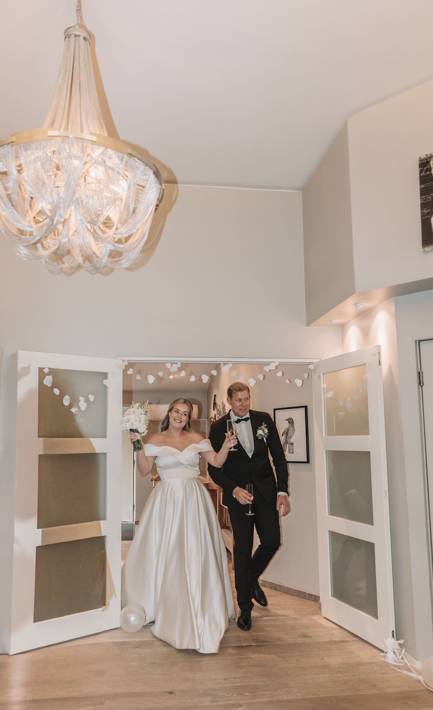 Bride in white off-shoulder gown and groom in black tuxedo making grand entrance through white French doors at wedding reception.