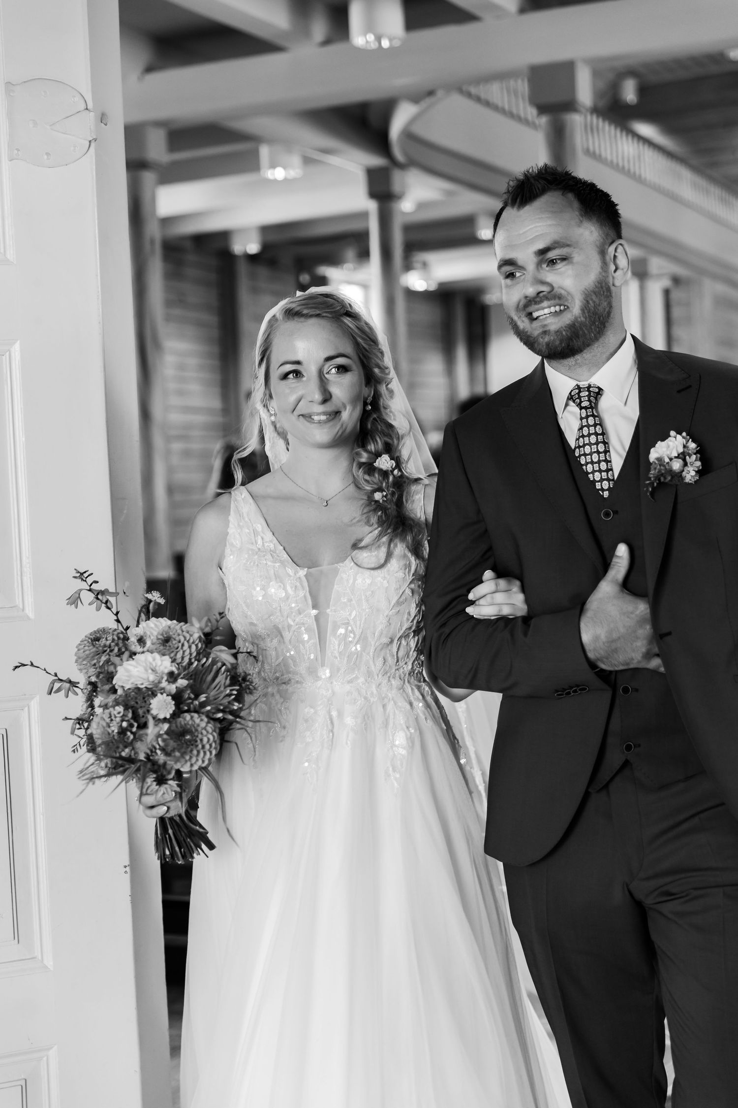 Black and white photo of a smiling bride holding a bouquet and groom in a suit walking together at their wedding.