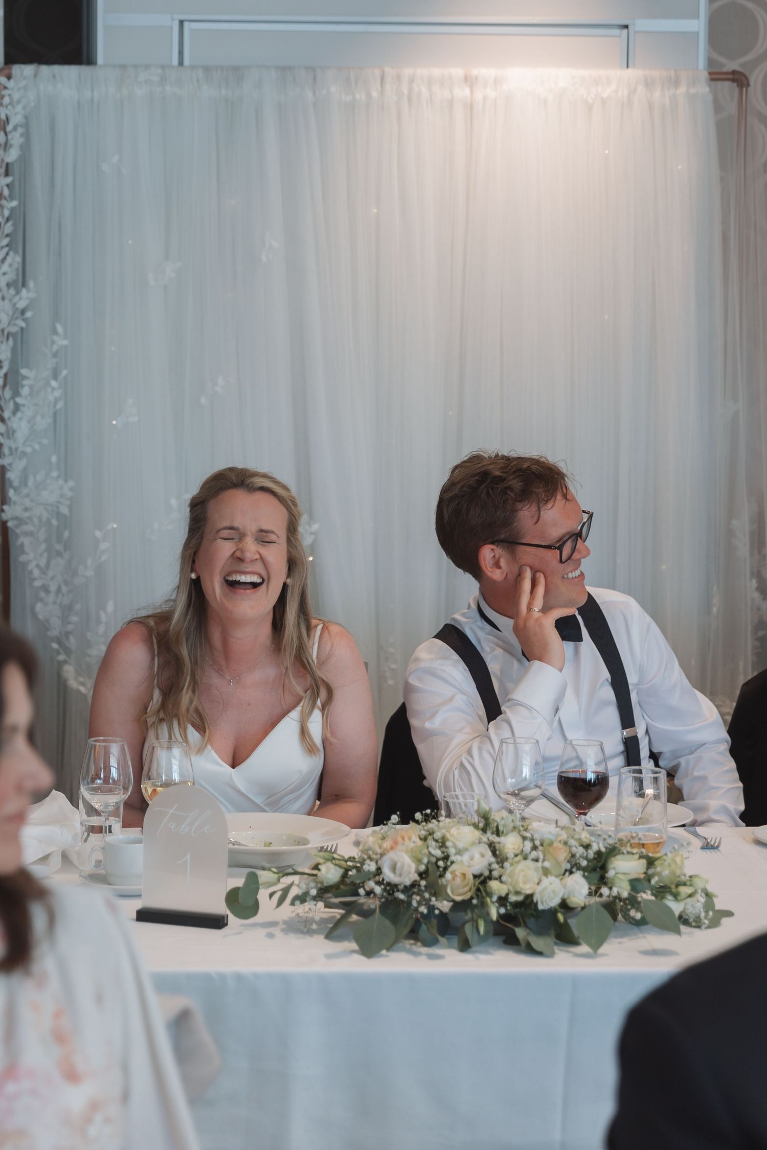 Bride laughing joyfully with groom at wedding reception head table adorned with white floral arrangements.