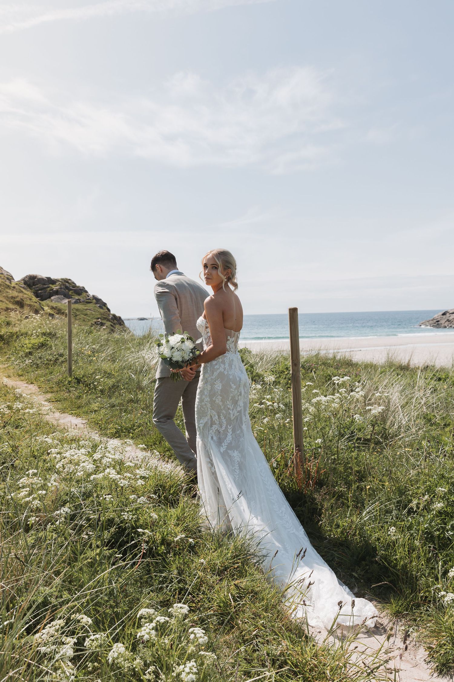 Bride in lace gown holds bouquet on coastal path, groom beside her, ocean and green hills in background.