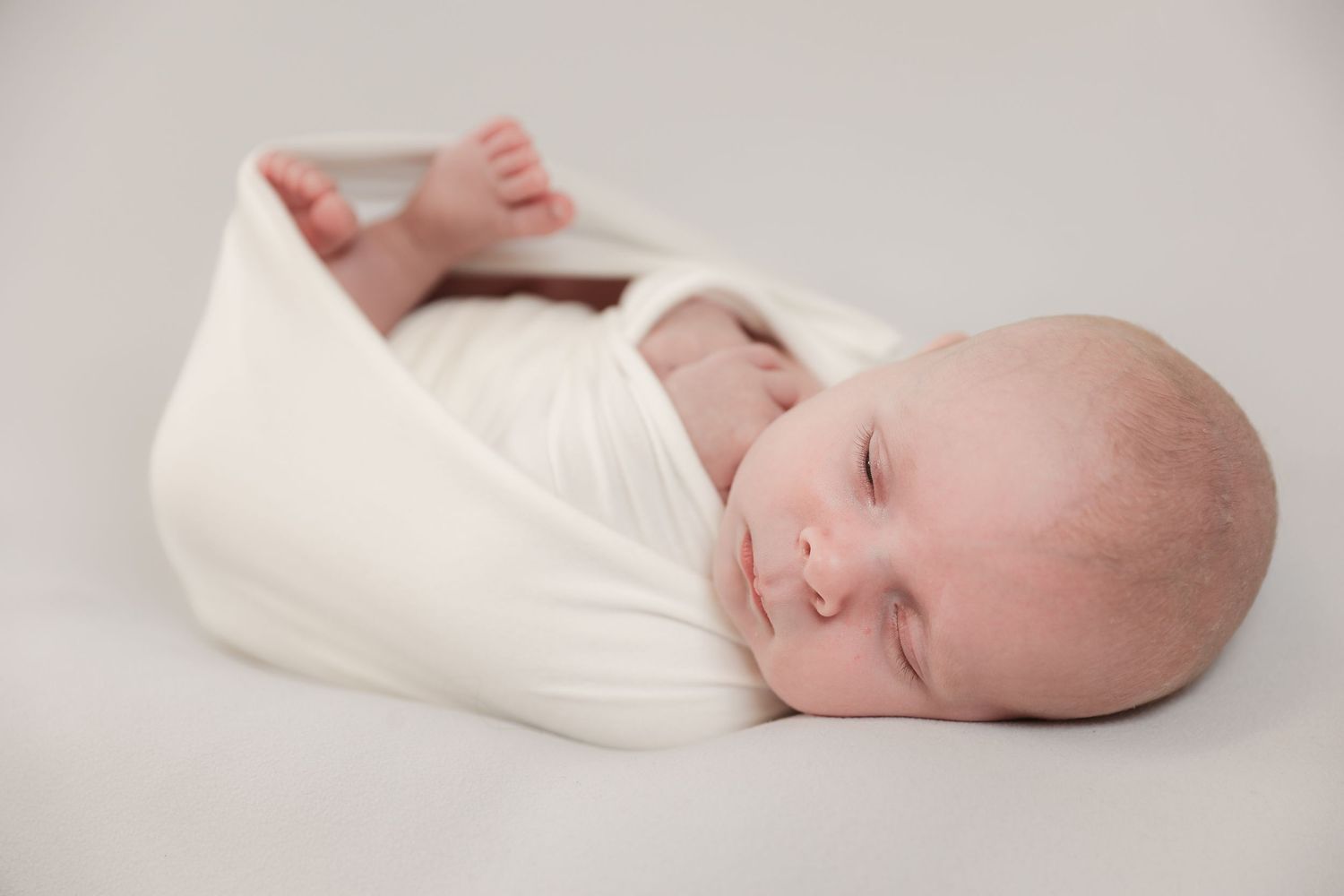 A newborn baby swaddled in white fabric lies peacefully in six different poses against a neutral backdrop.