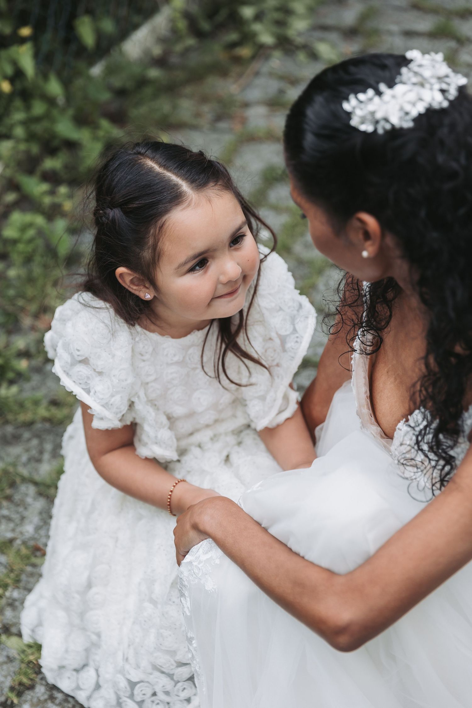 A bride in a white gown holds a young flower girl in a lace dress, sharing a sweet moment outdoors.